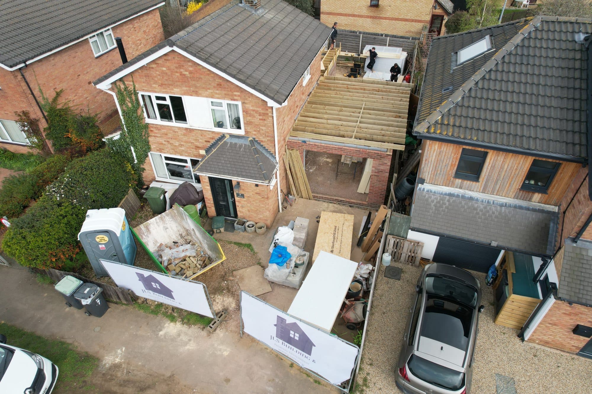 Aerial view of the rear extension and garage under construction, with new timber roof joists, a skip and JLS Building site banners in the driveway, All Saints Way, Sandy