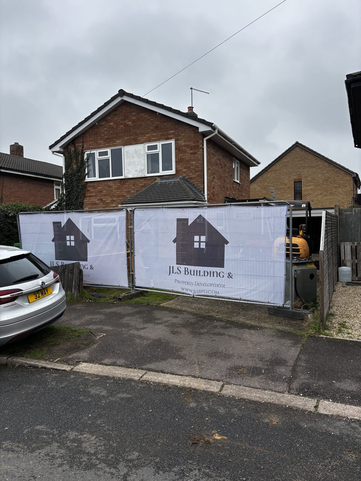 Site hoarding with JLS Building & Property Development banners strung across the driveway in front of the brick detached house