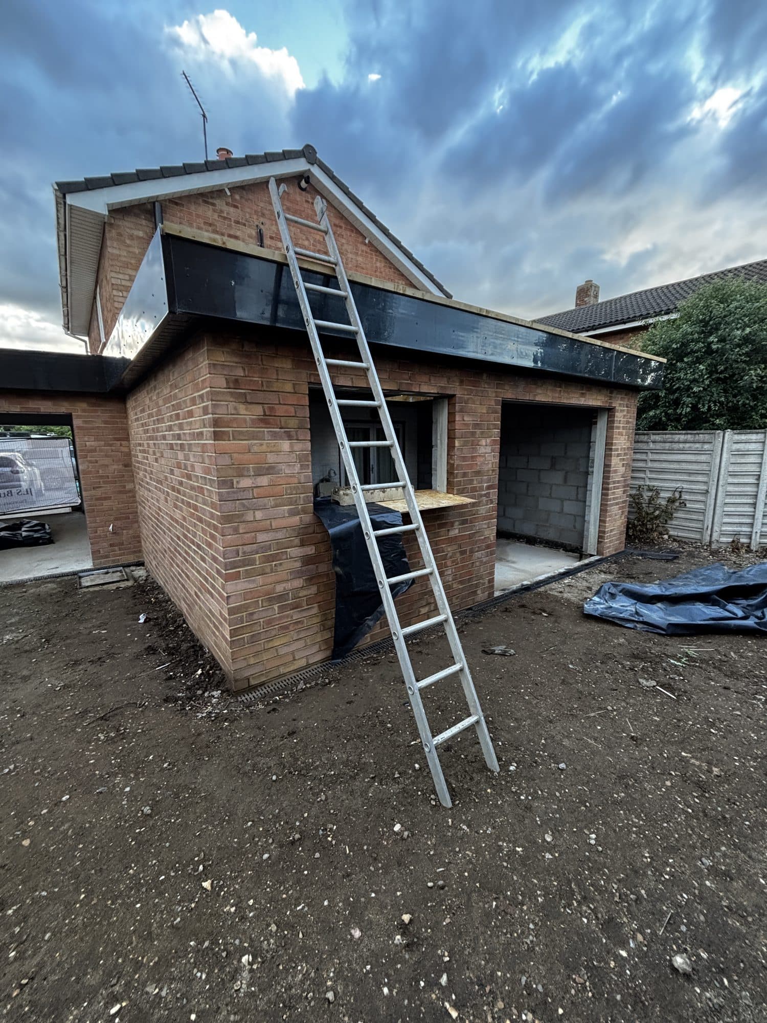 Ladder leaning against the brick extension and garage walls with black roof edge trim installed, under a cloudy sky