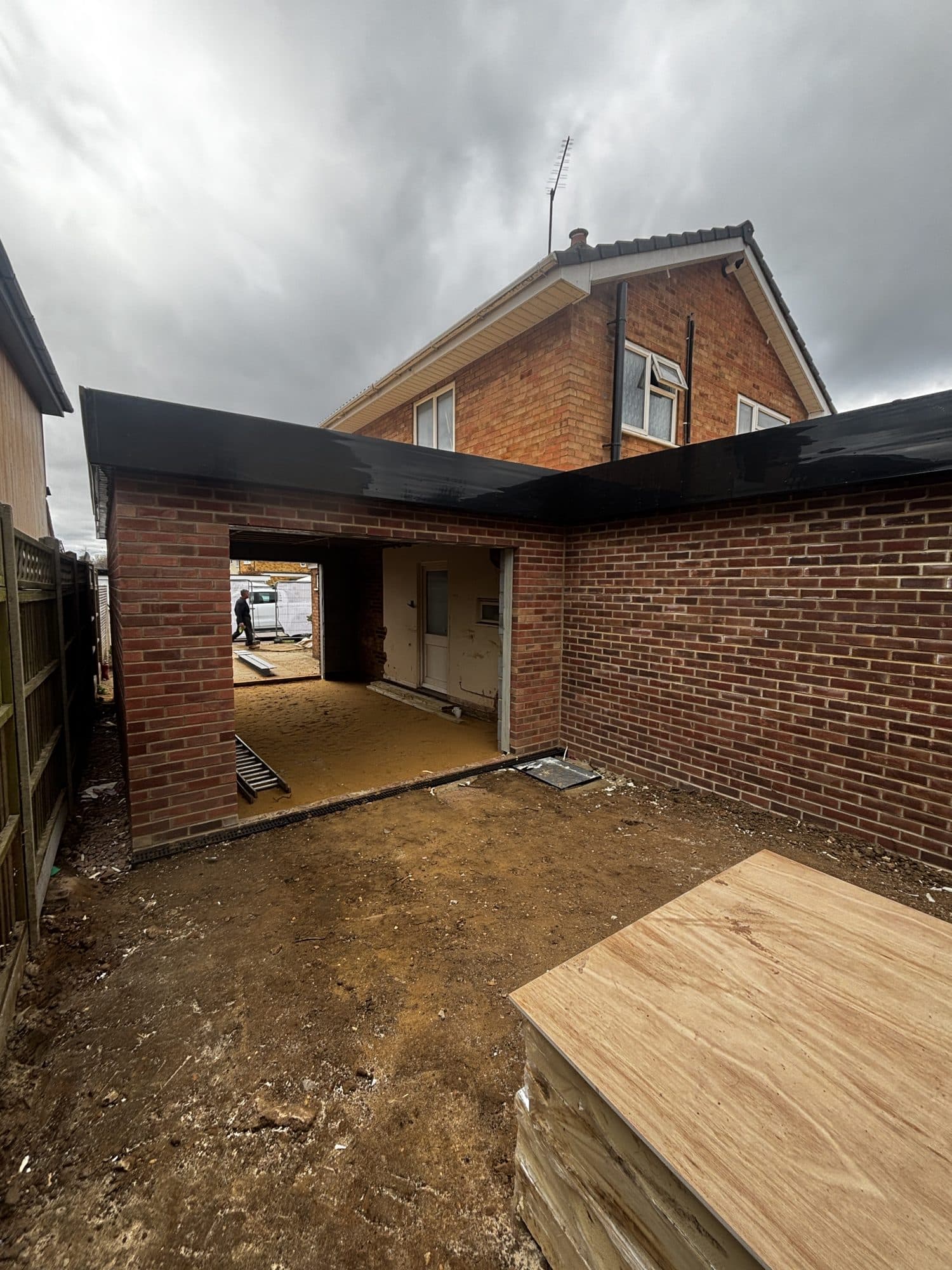 Rear view of the brick extension and garage shell with black roof trim, an open doorway through to the house and plywood sheets stacked outside