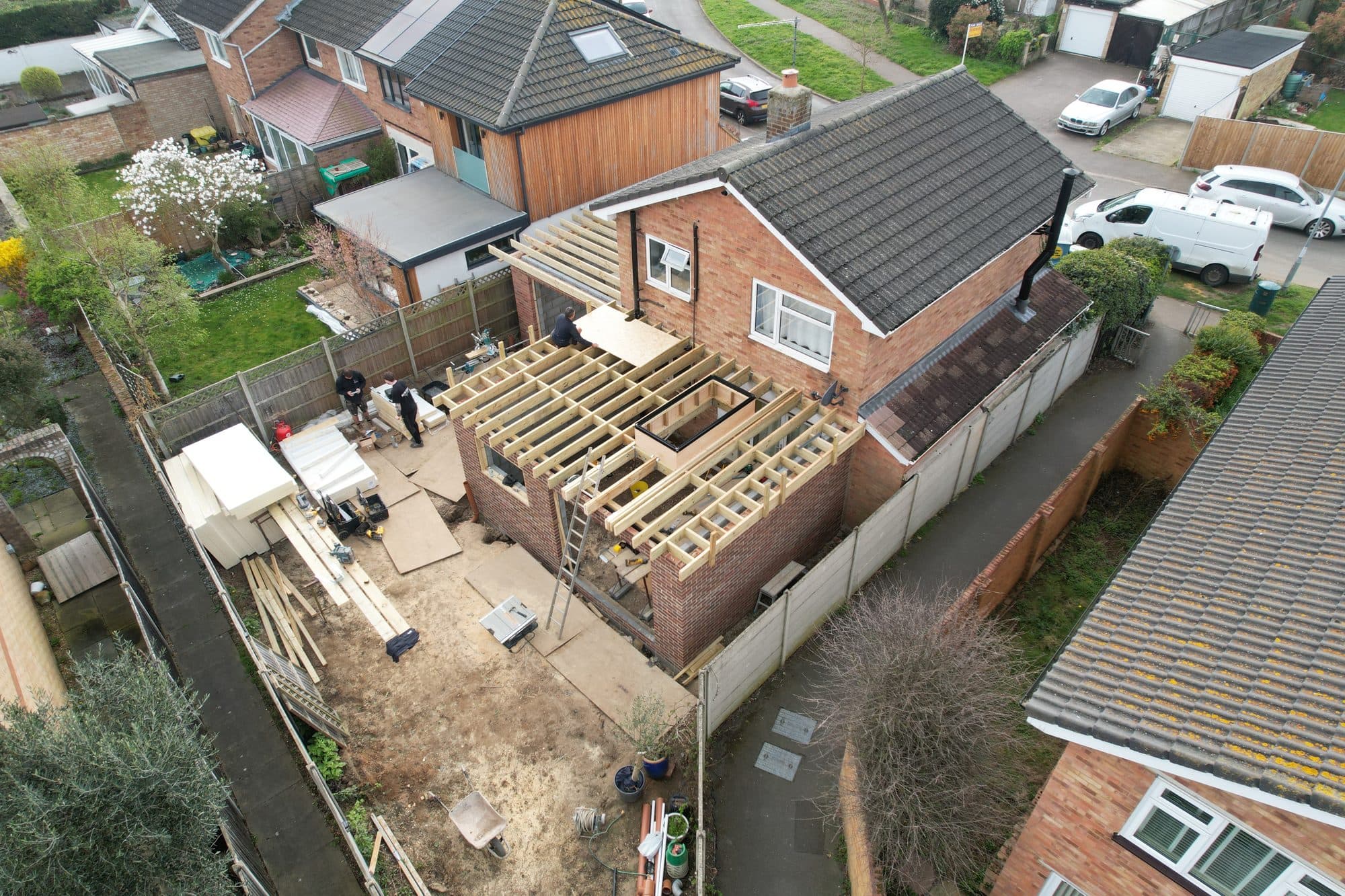 Aerial view of the extension roof under construction with timber joists, part OSB boarding, two workers fitting panels, and insulation boards stacked in the garden below