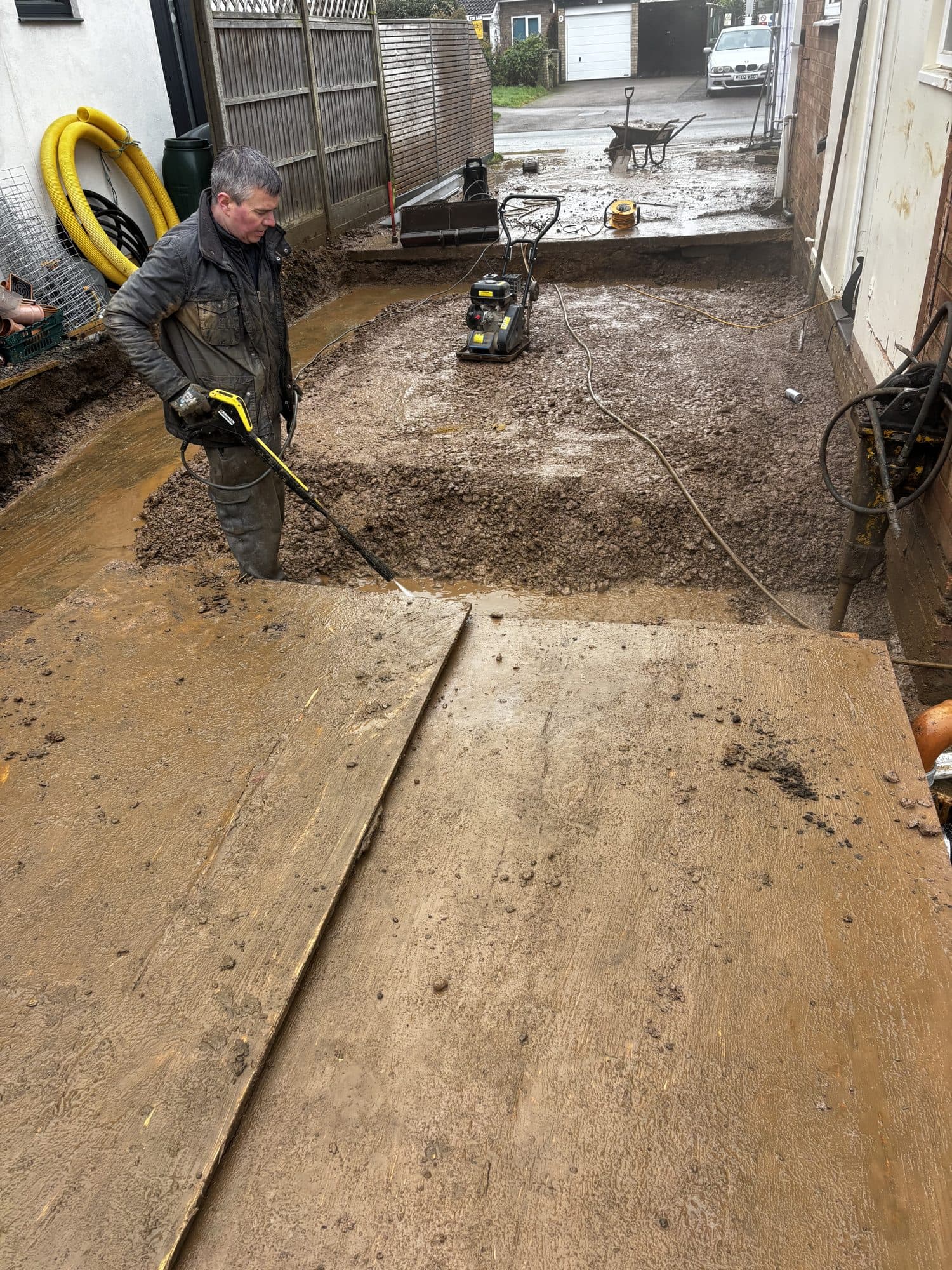 Worker pressure washing a freshly laid stone sub-base in the garden extension area, with a plate compactor and wheelbarrow nearby