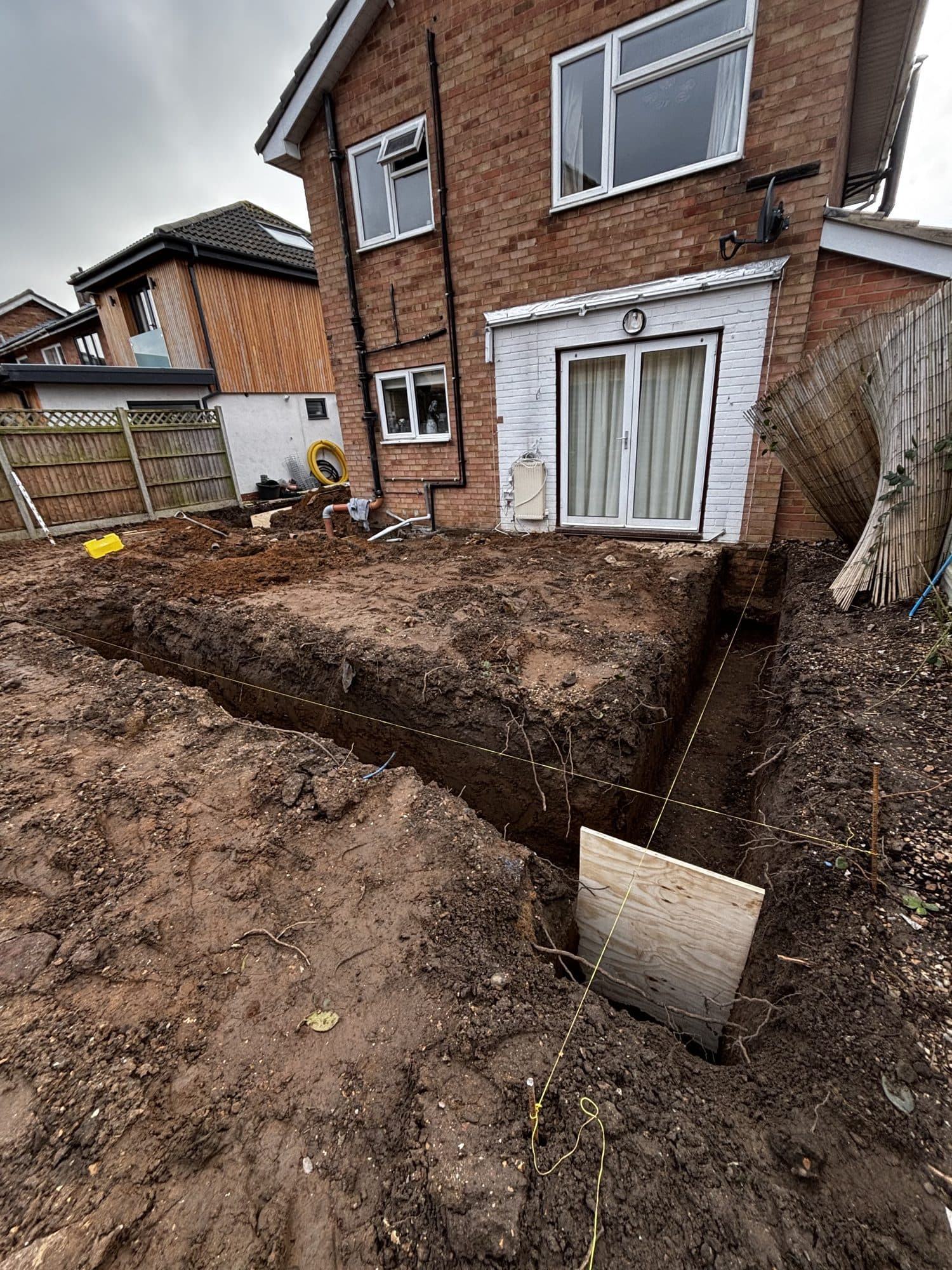 Foundation trenches dug out in the rear garden with string lines marking the extension layout, two-storey brick house behind