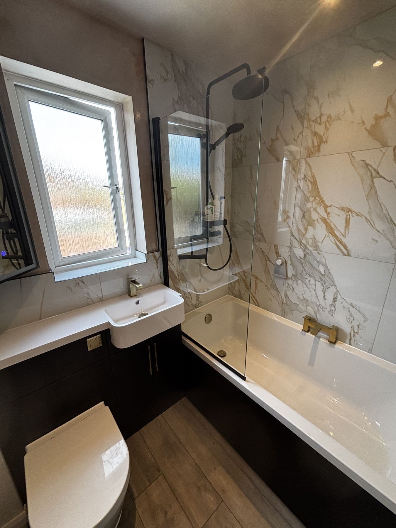 Bath with glass shower screen, marble tiled walls and black vanity unit beneath a window