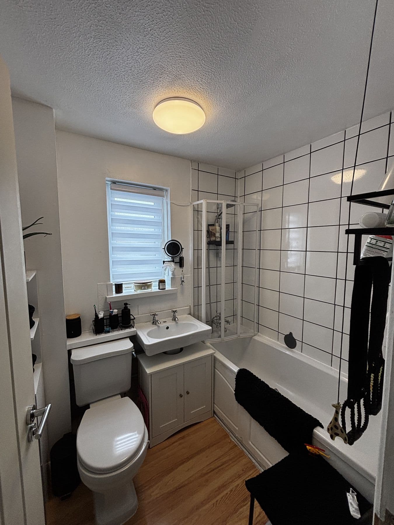 Older bathroom with white tiled walls, folding shower screen, wall hung sink and toilet beneath a window