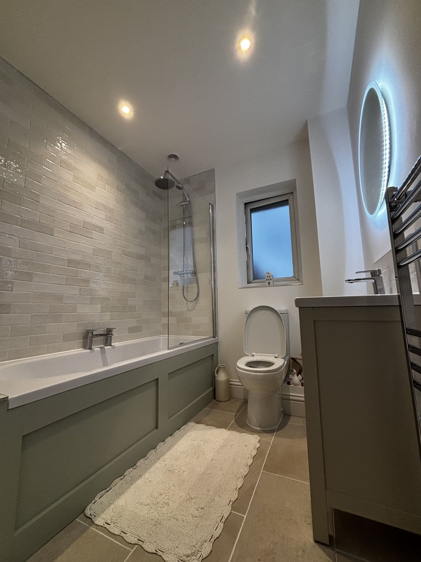 Finished bathroom with bath and glass shower screen, grey brick-effect tiling, green panelled bath front, wall-hung toilet and illuminated round mirror above a vanity basin, Peer Road, St Neots