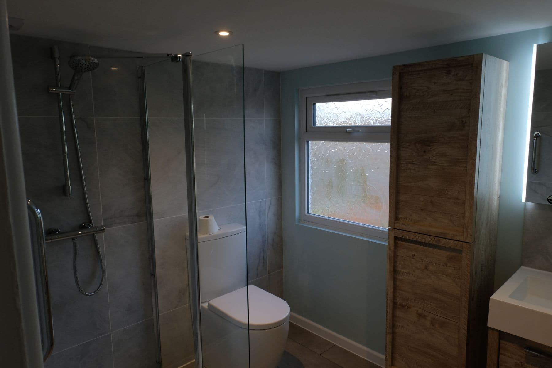 Shower room with walk-in glass shower screen, grey tiling and wood effect tall cabinet, Cotton End