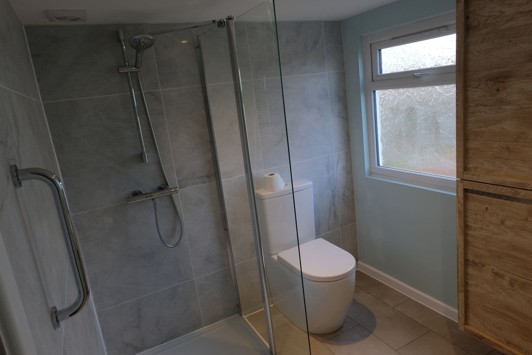 Walk-in shower with chrome riser rail and handset against grey tiled walls, toilet beside the window