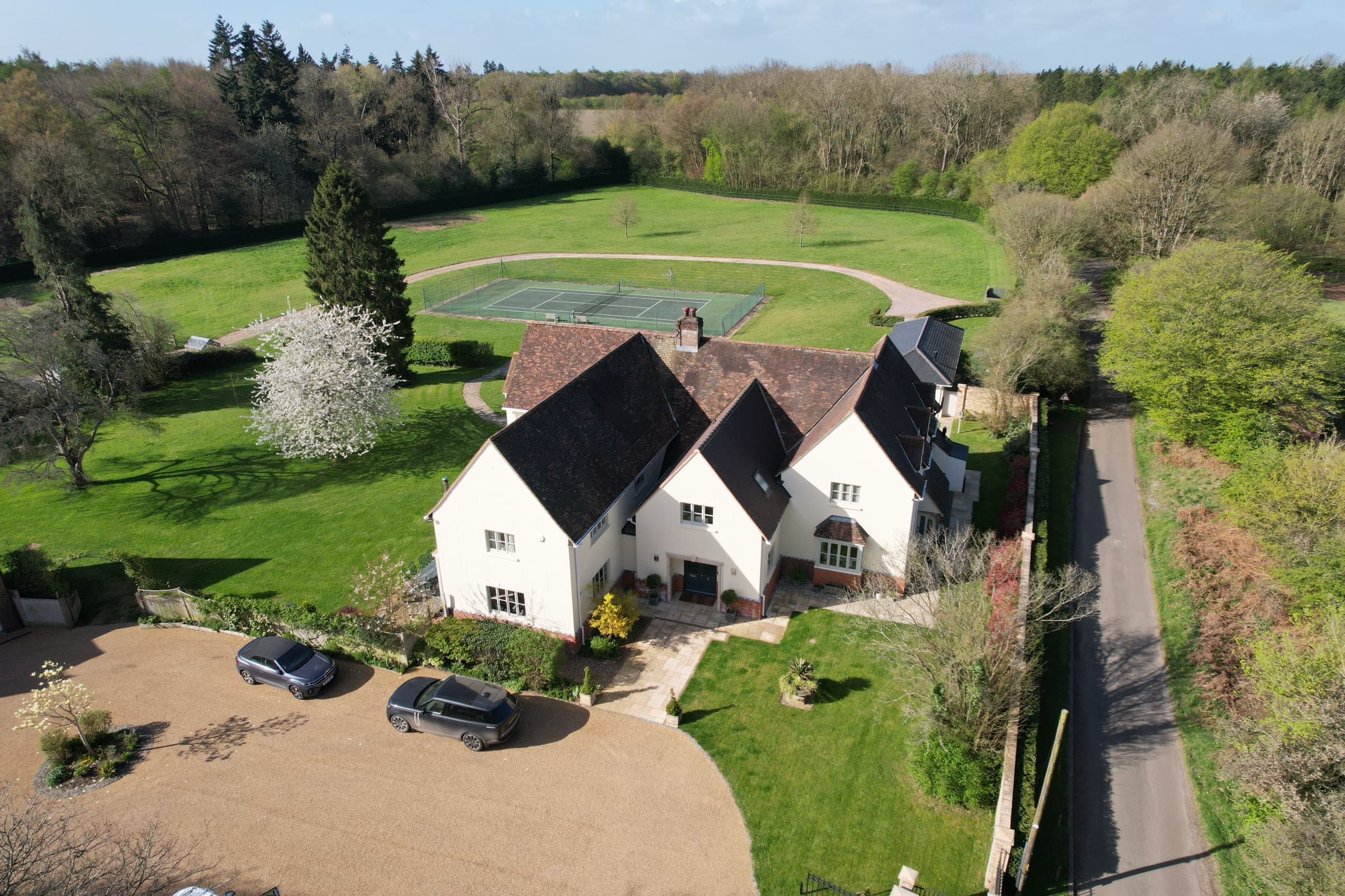 Aerial view of the house, tennis court and gardens from the driveway side with two cars parked