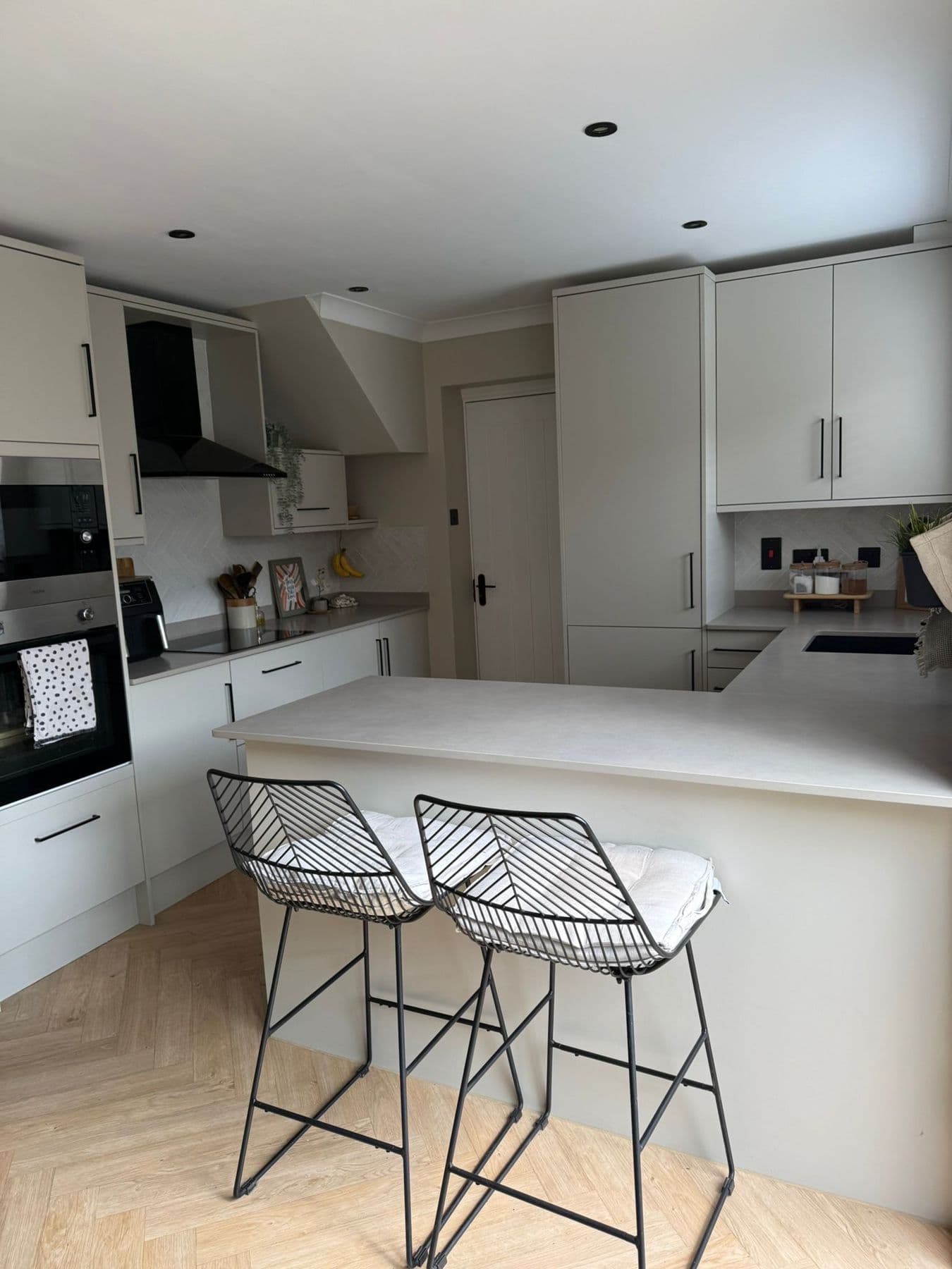 Open U-shaped kitchen in light grey cabinetry with a central breakfast bar, black wire frame stools and oak effect flooring, Falcon Close, Sandy