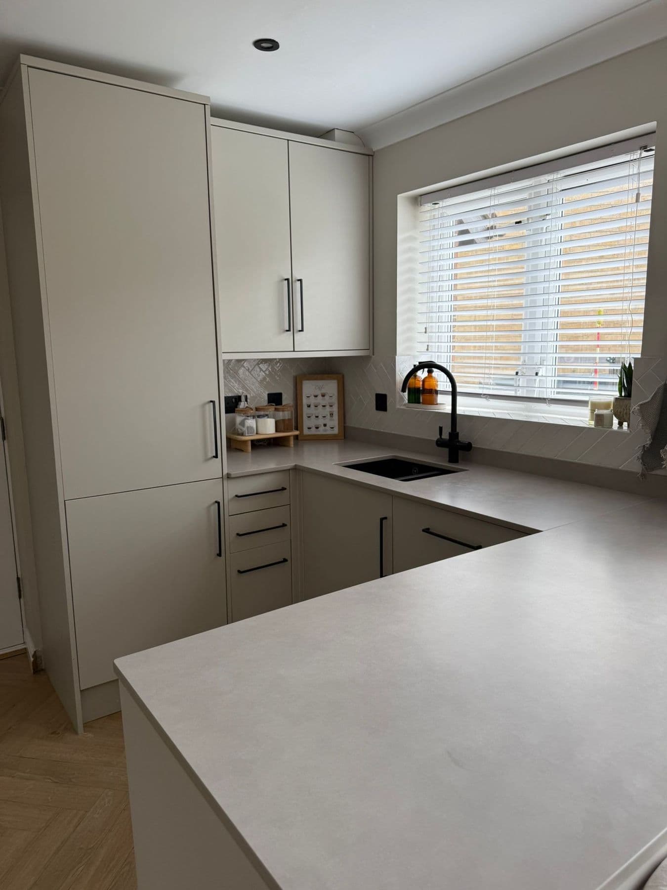 Light grey U-shaped kitchen with a black mixer tap over an undermounted sink, grey worktop and a window with white venetian blinds