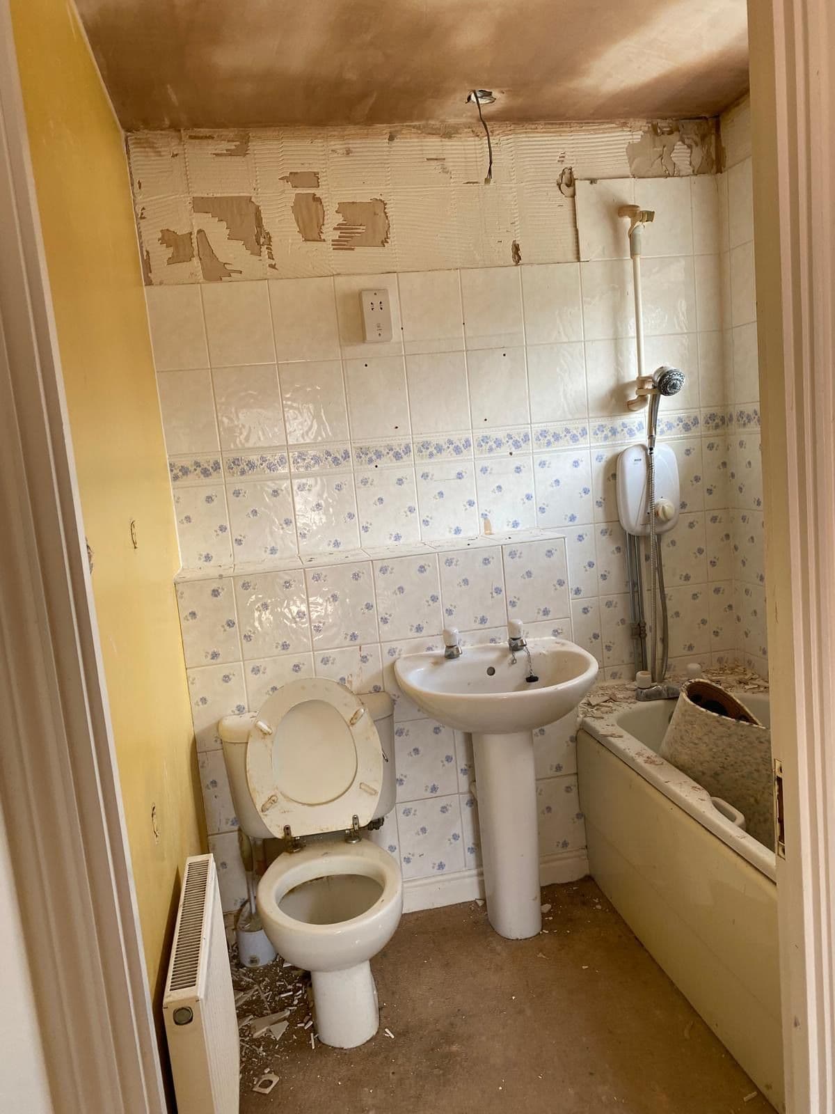 Bathroom mid strip out with partially removed wall tiling, an old pedestal basin, toilet and bath before renovation