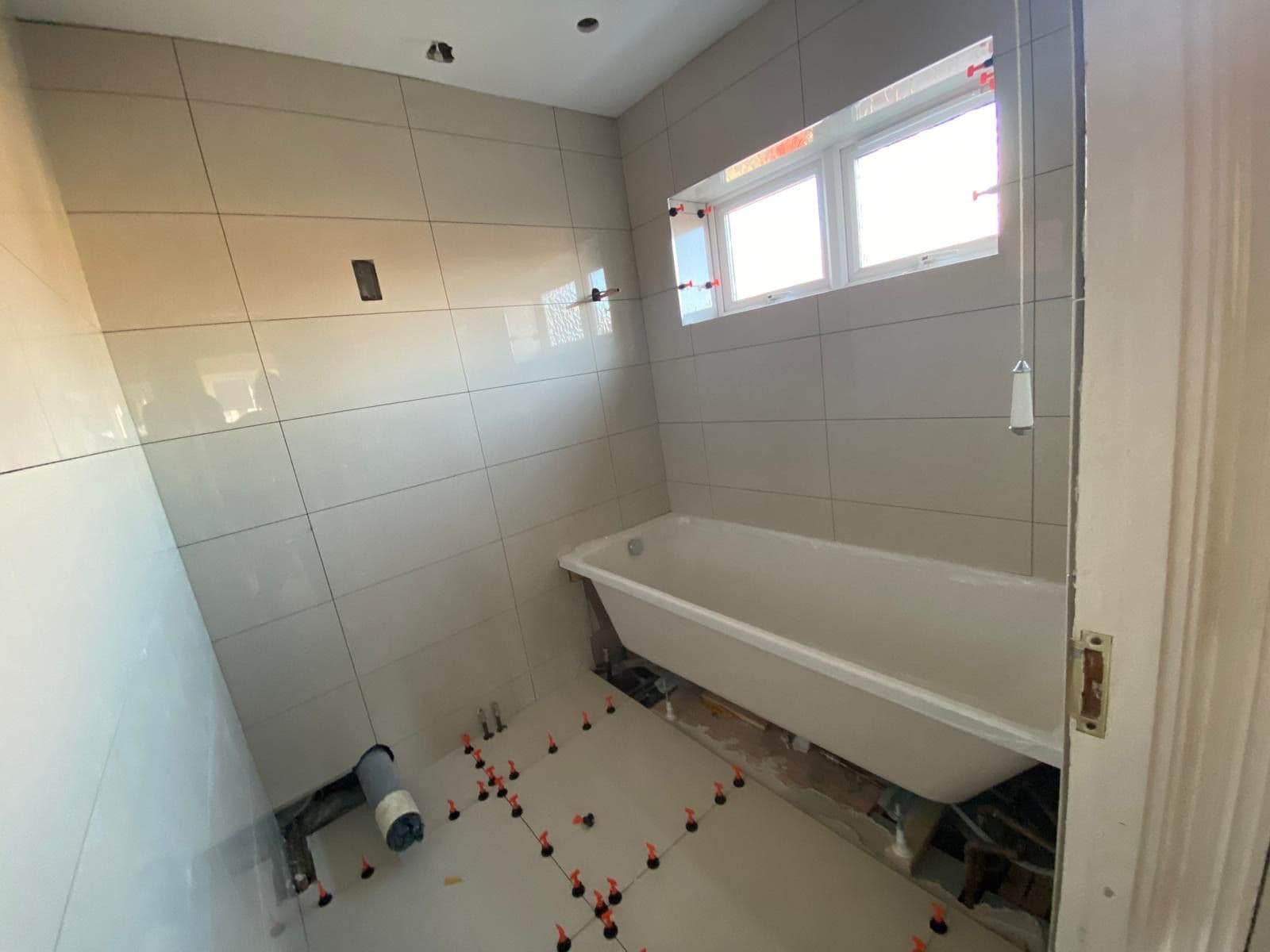Bathroom in progress with new floor tiles being laid using tile spacers, a white bath installed and a window above