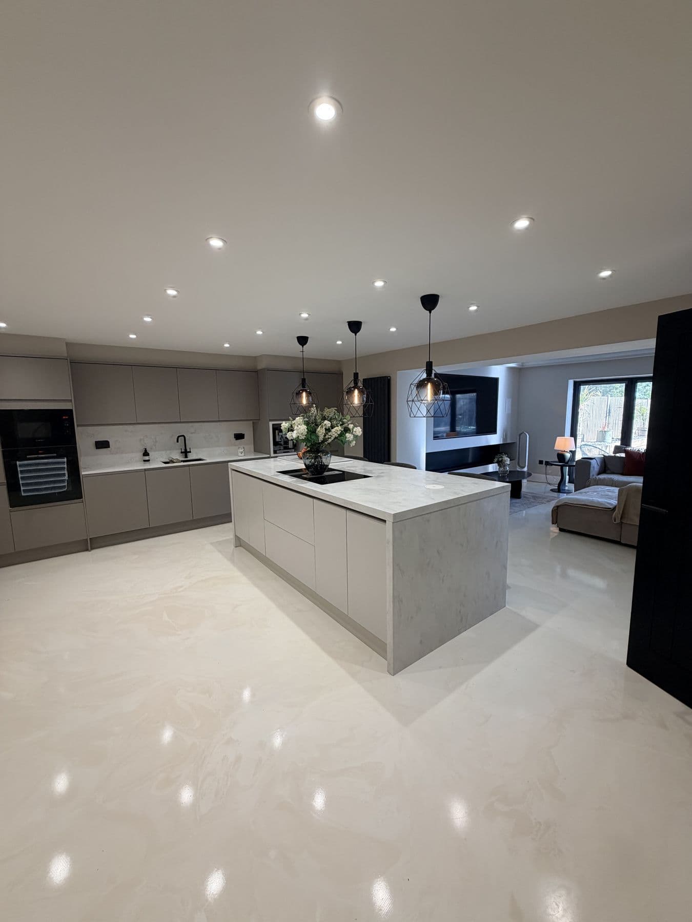 Kitchen island with white marble effect worktop and waterfall end panel, black wire pendant lights, opening onto a living area with sofa and wall mounted TV