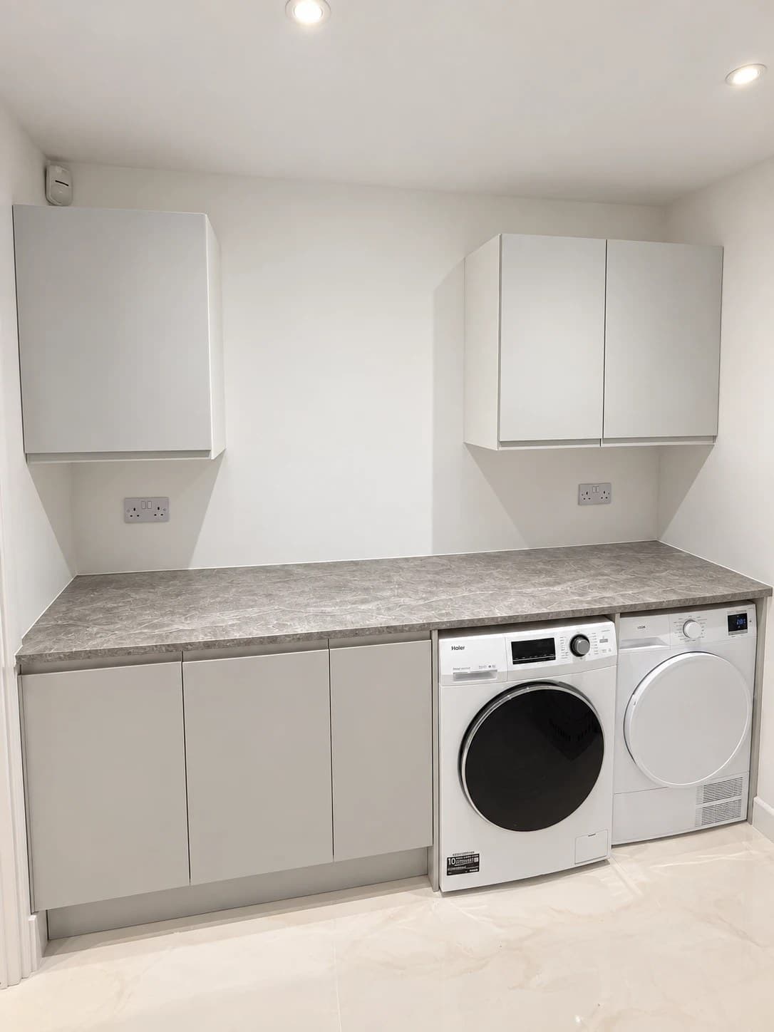 Utility room with light grey wall and base cabinets, a grey marble effect worktop and a white washing machine and tumble dryer side by side