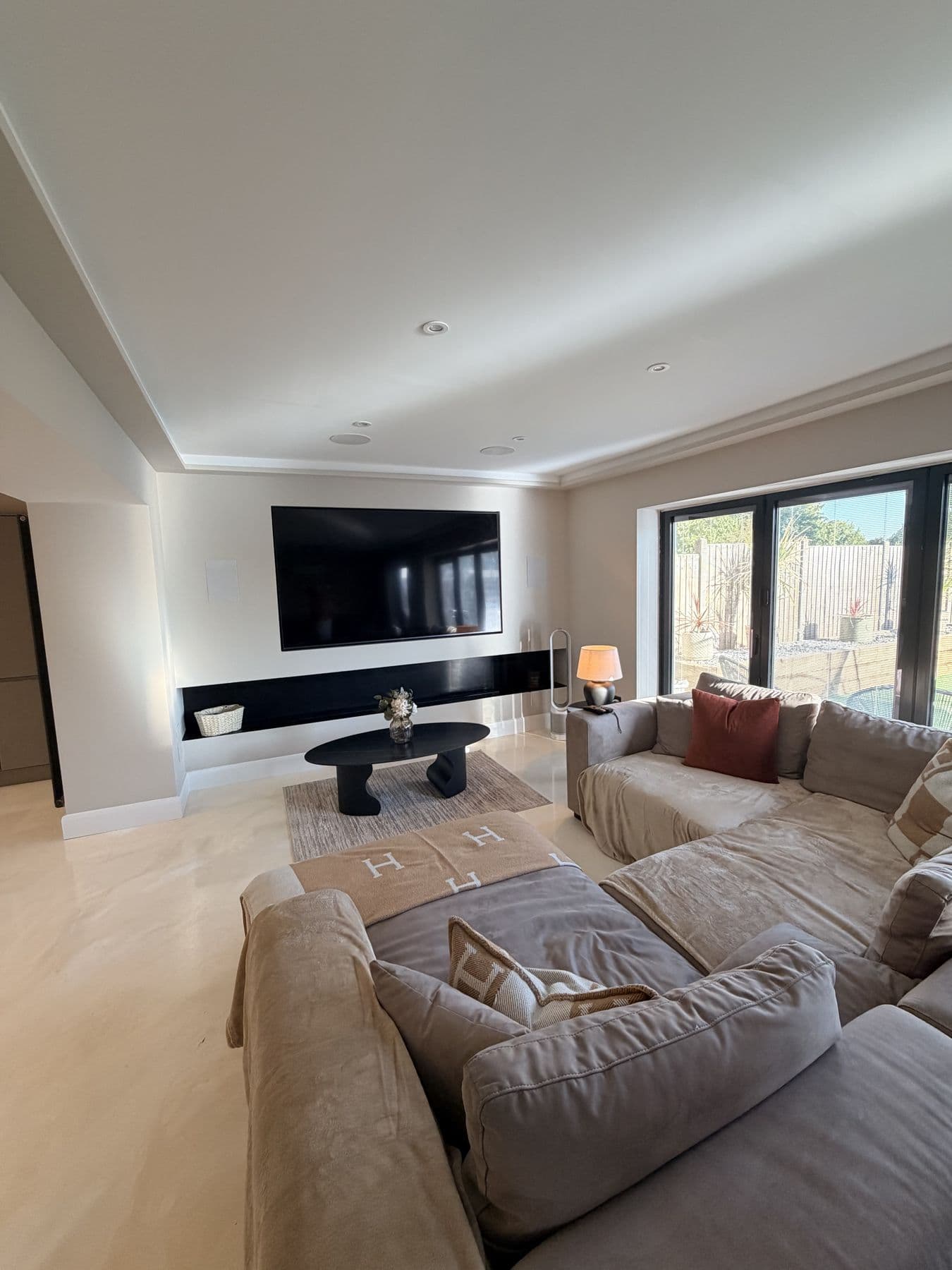 Living room with an L-shaped grey sofa, black oval coffee table, wall mounted TV and bifold doors onto the garden