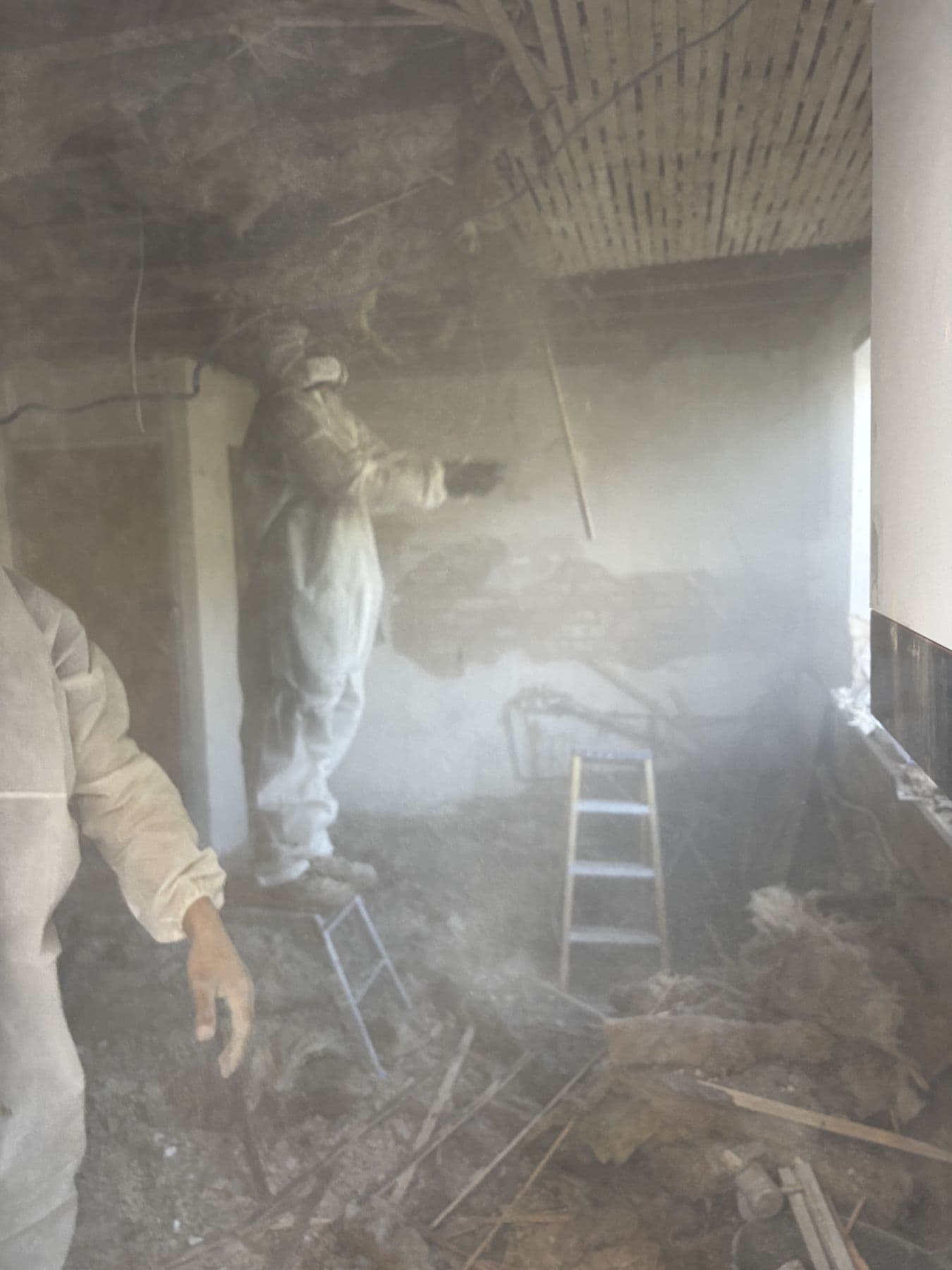 Worker in white protective coveralls amid thick dust during ceiling demolition, insulation and timber debris covering the floor