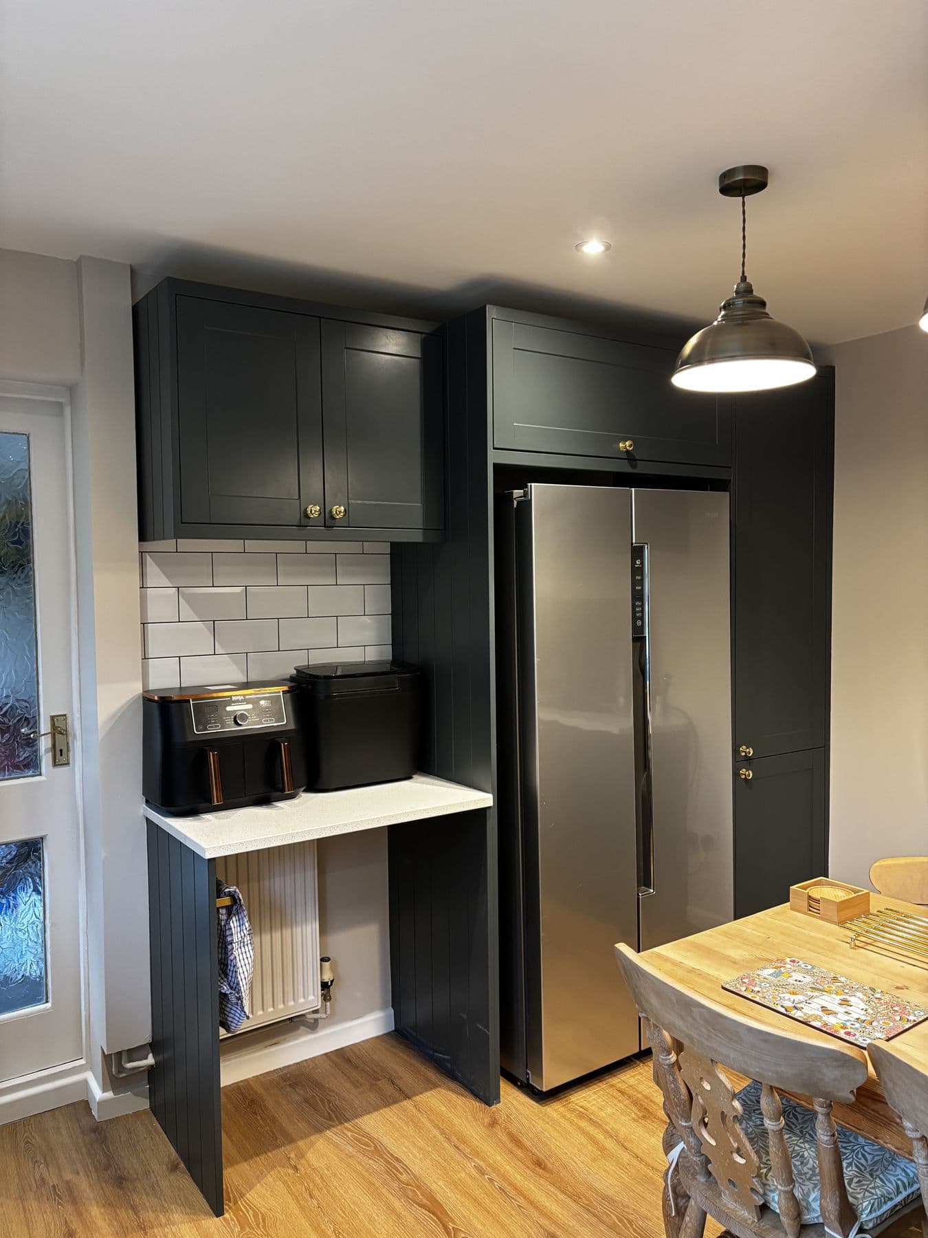 Dark green kitchen cabinetry around a stainless steel American style fridge freezer, with a pendant light over a wooden dining table