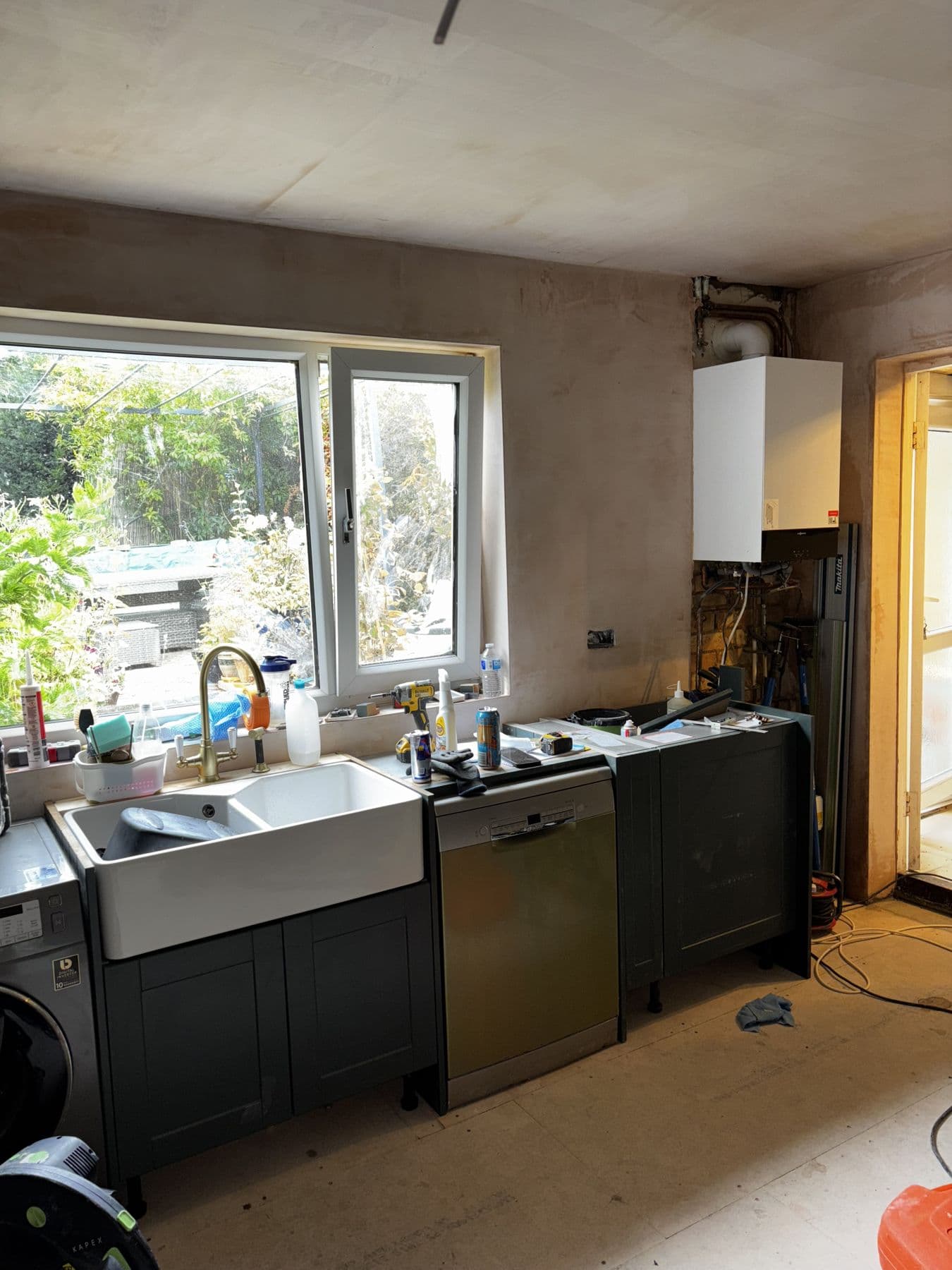 Kitchen mid renovation with freshly plastered walls, a wall mounted boiler, sink unit and dishwasher installed but worktops and flooring not yet fitted