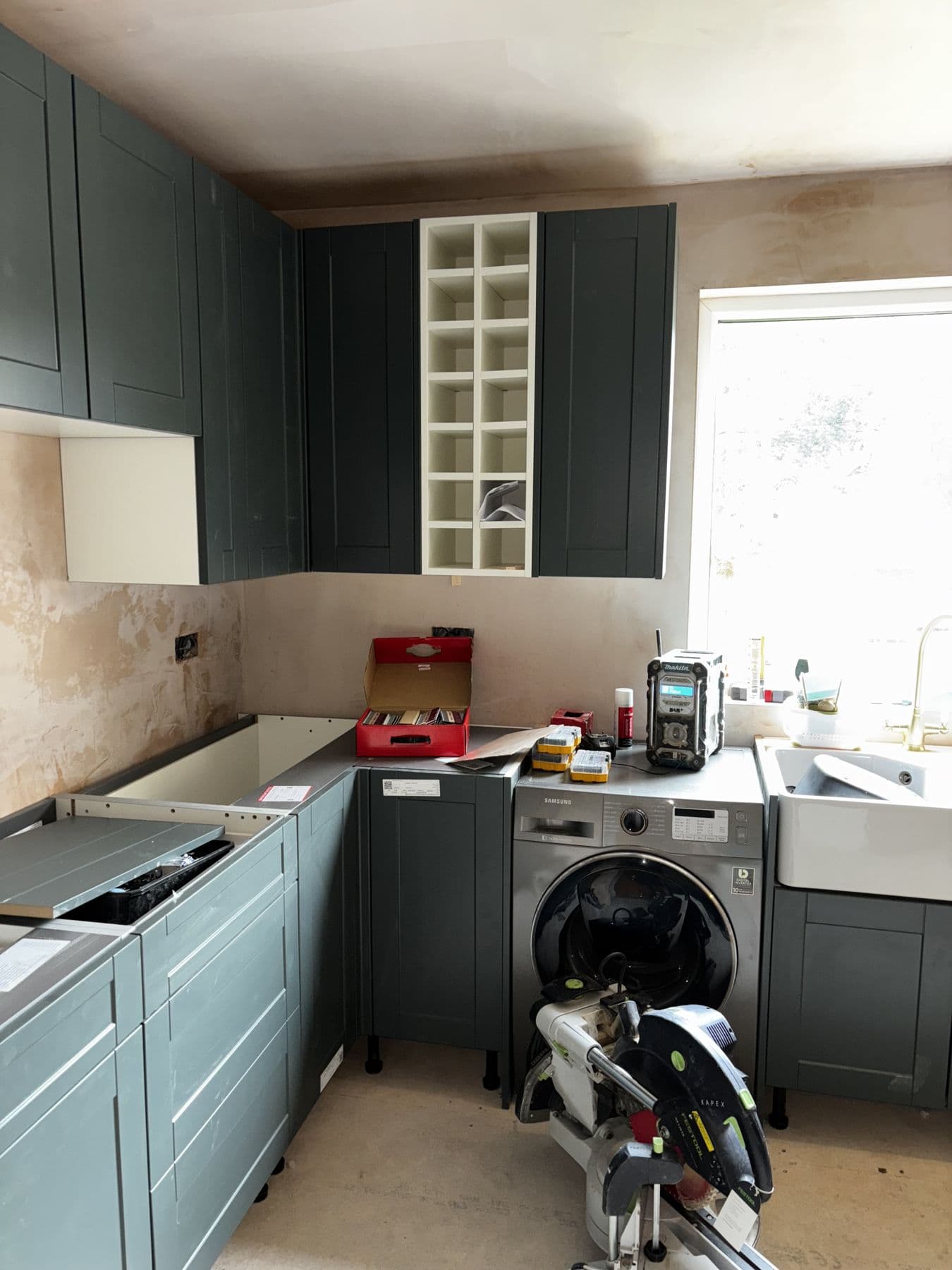 Kitchen cabinets being installed, including a dark green wall unit with a built-in wine rack, plastered walls still bare and tools left on the worktop