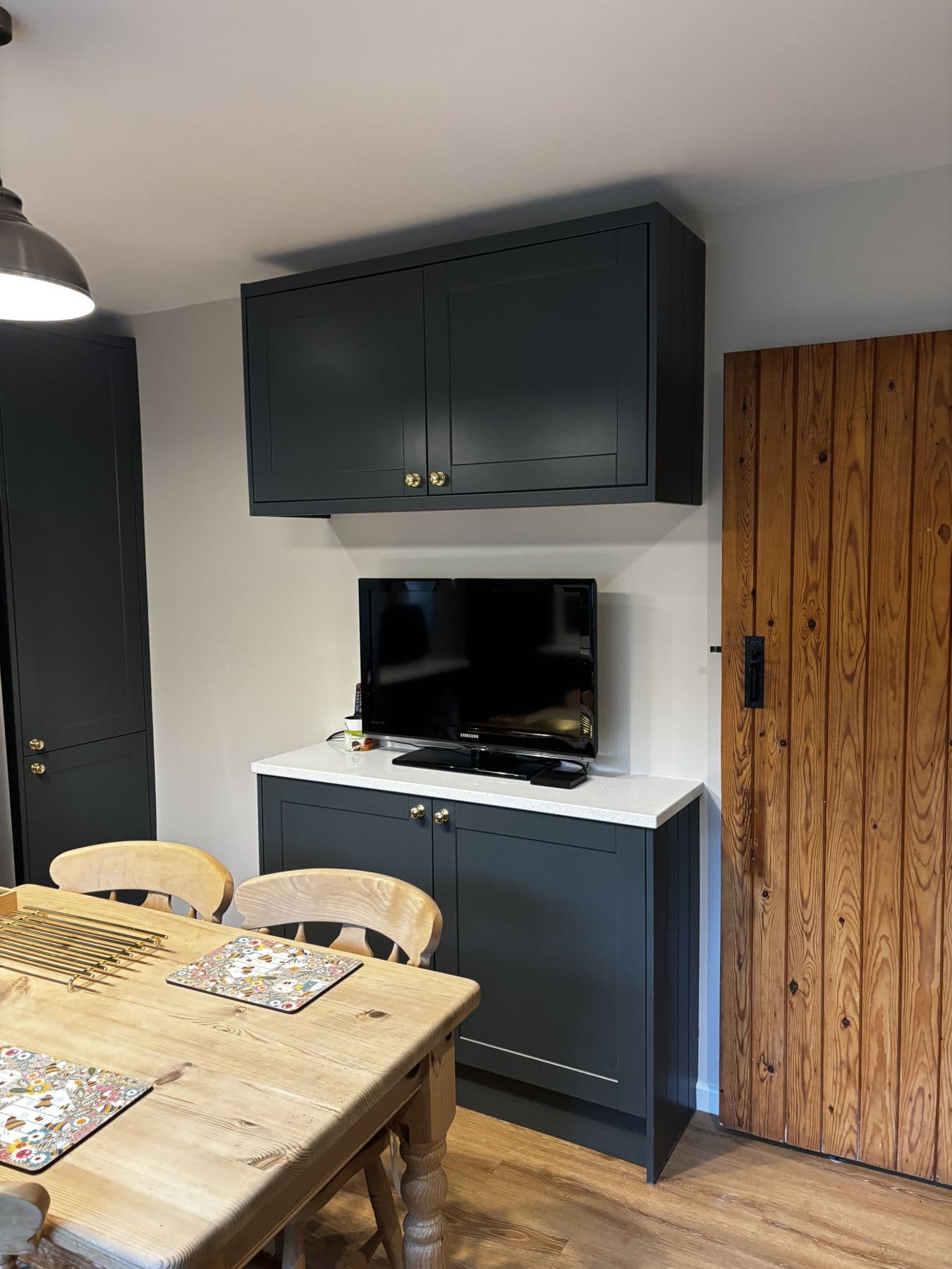Dark green wall cupboard above a wall mounted television and sideboard, next to a panelled wooden door in the finished kitchen diner