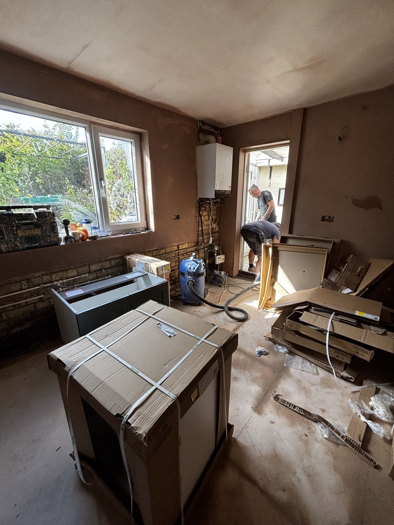 Kitchen renovation in progress with flat pack cabinet carcasses and a boxed appliance on the floor, exposed brickwork and two workmen at the back door