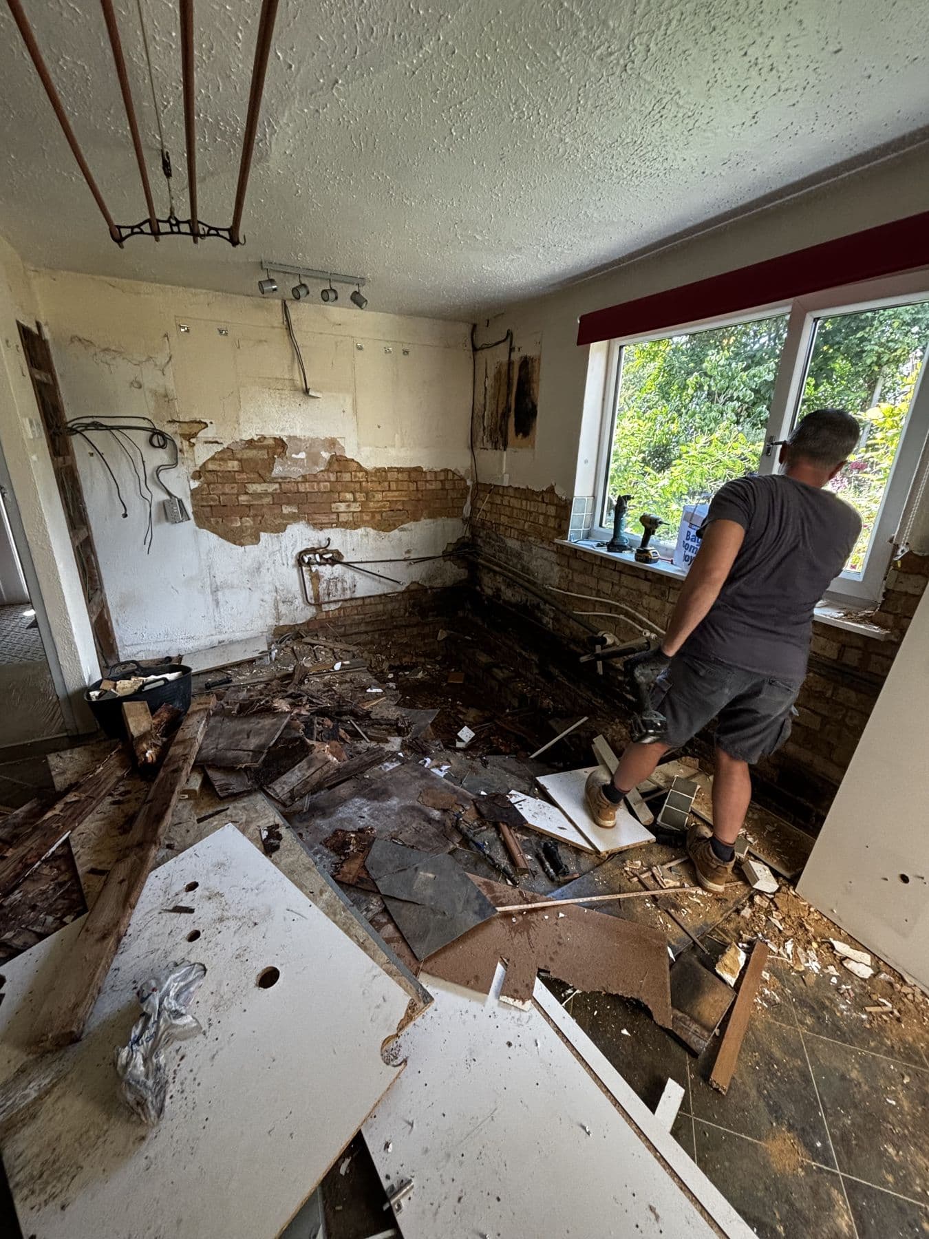 Kitchen mid strip out with broken floor tiles and rubble scattered across the room and a worker clearing debris near the window