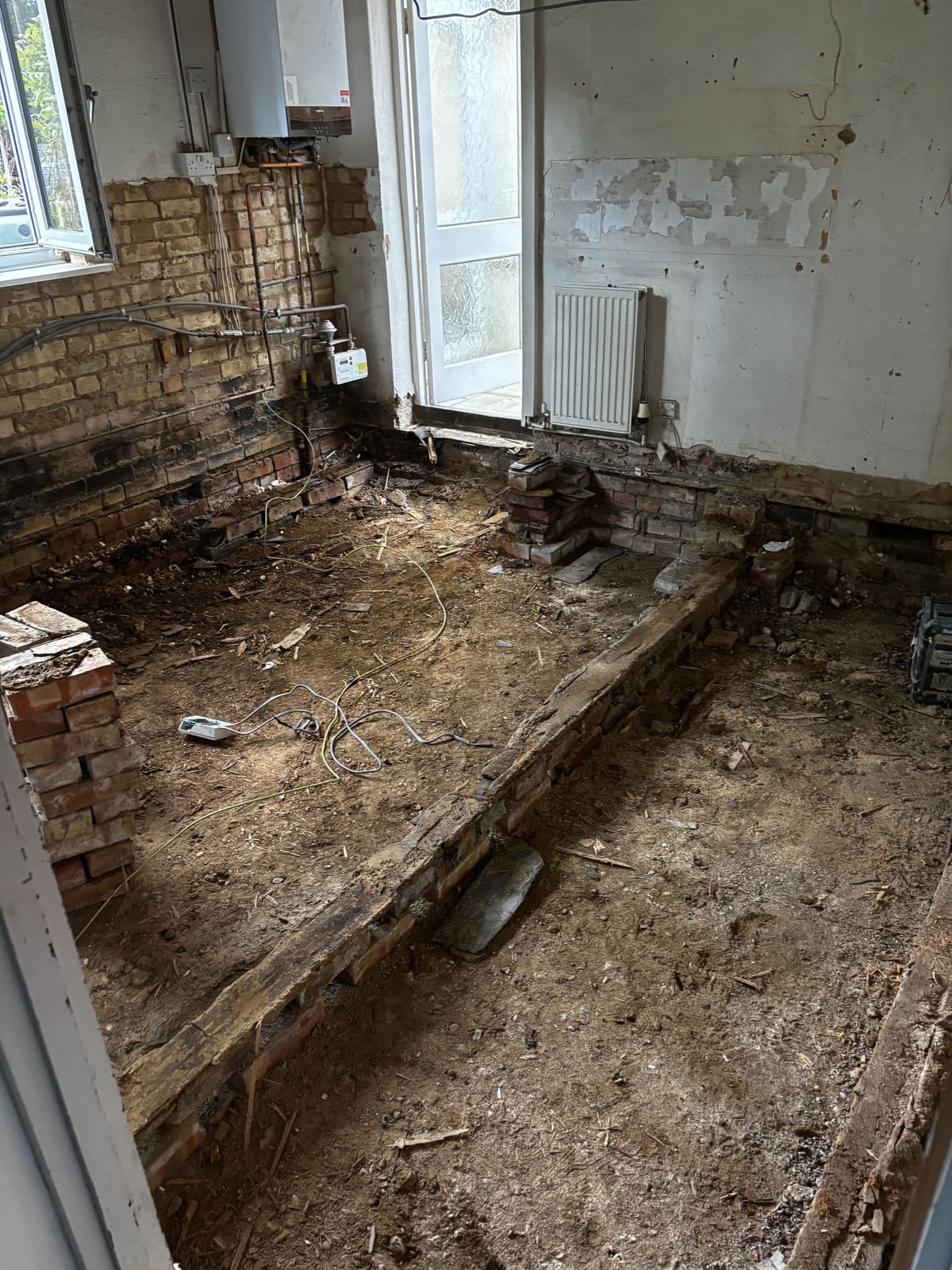 Old suspended timber floor removed down to bare earth and brick sleeper walls, exposed pipework and a radiator against the wall
