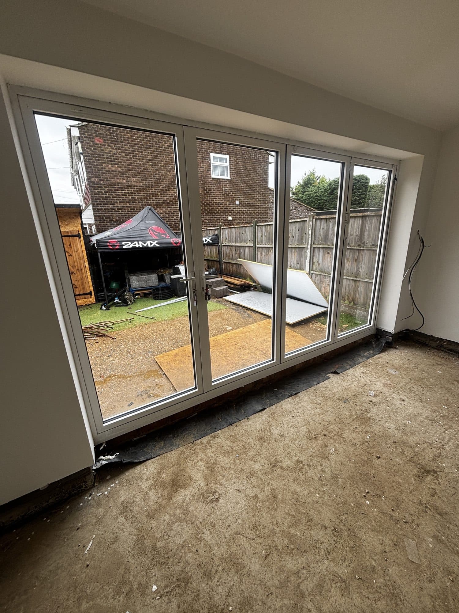 Interior view through newly fitted white aluminium bifold doors onto the garden, extension still on a bare concrete subfloor