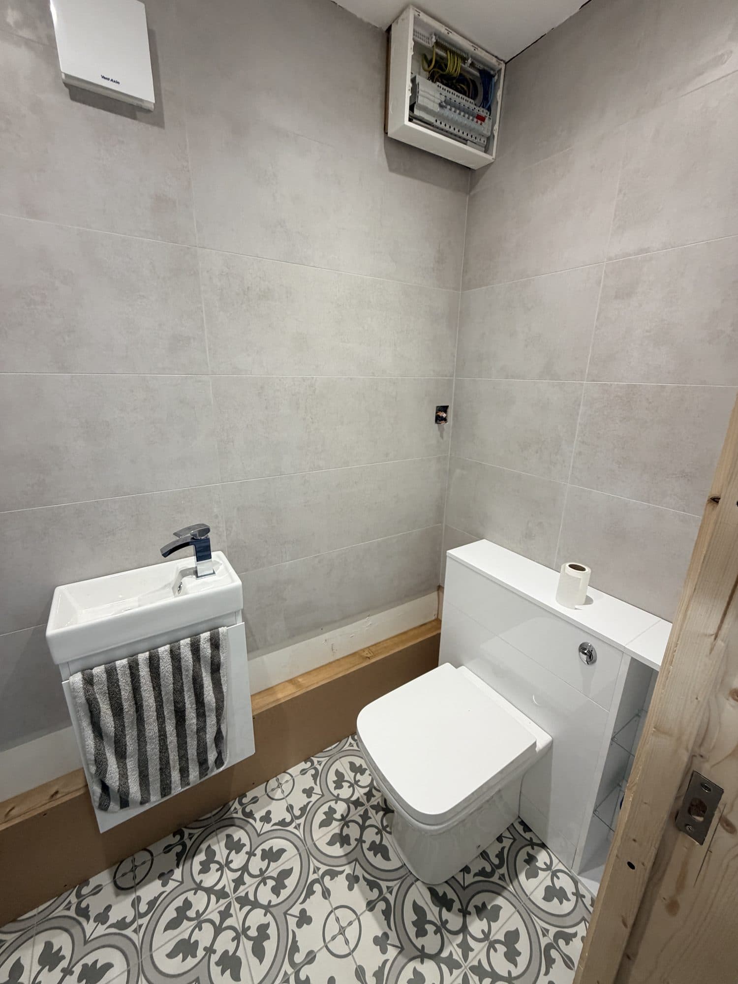 Downstairs toilet under construction, with patterned tiled floor, a small basin and toilet fitted, and an exposed electrical consumer unit on the wall