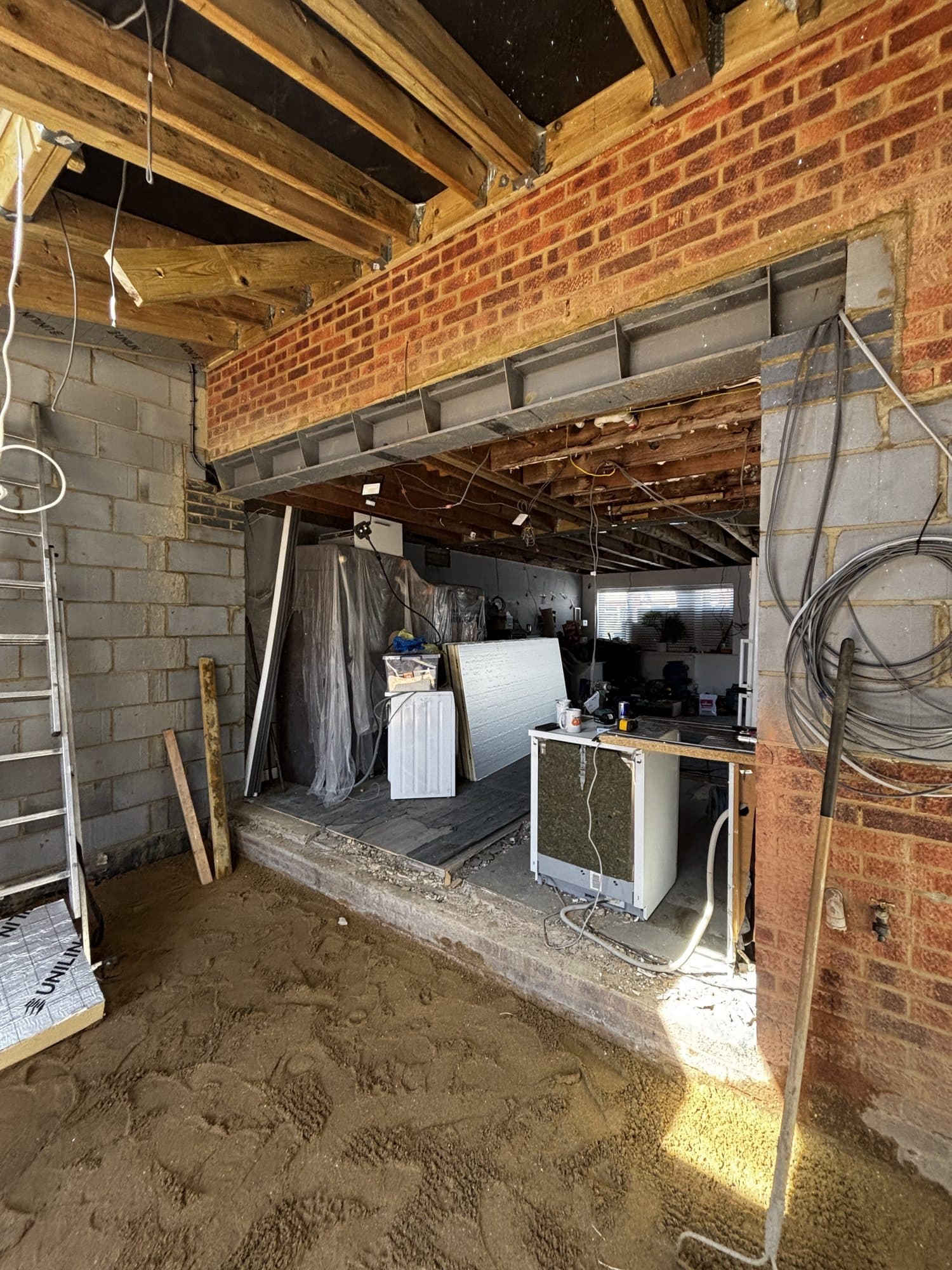 Inside the extension mid build, showing exposed timber floor joists, a steel lintel over the new opening and blockwork walls