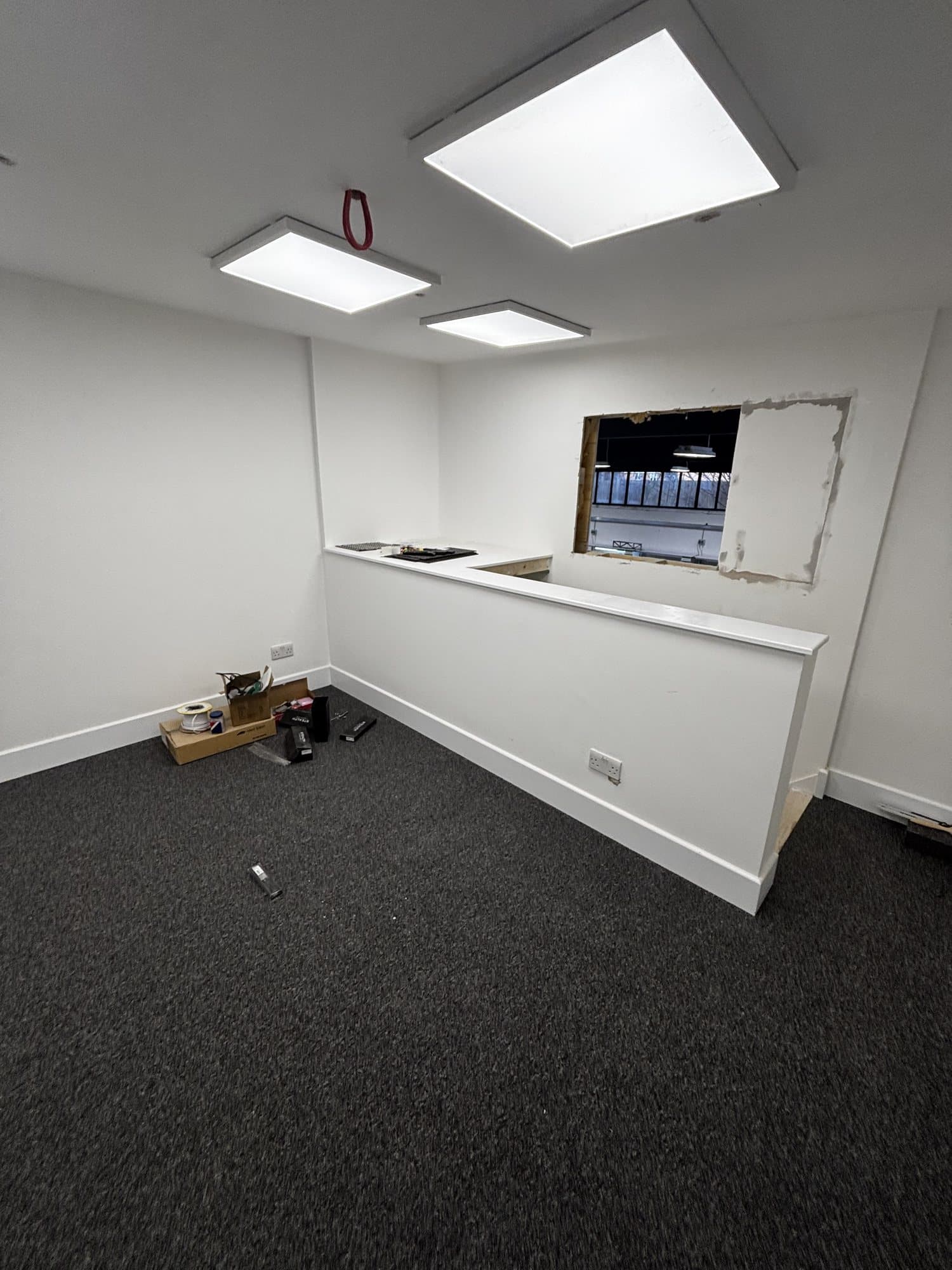 Room with a built in reception counter, carpet and ceiling lights, with an unfinished square cut out in the wall for a window