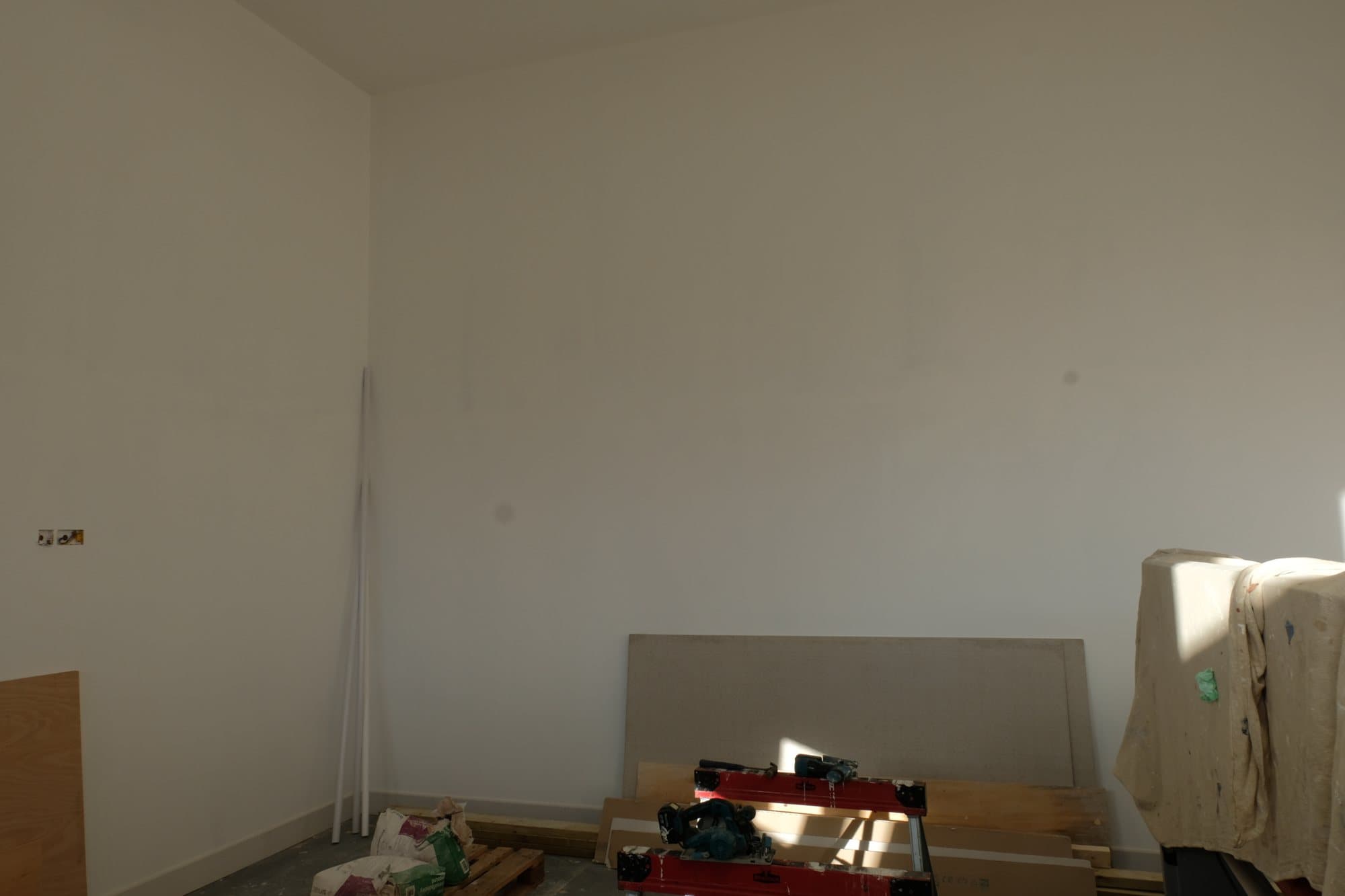 Plastered room with bare walls and construction materials including plasterboard and power tools stacked on the floor