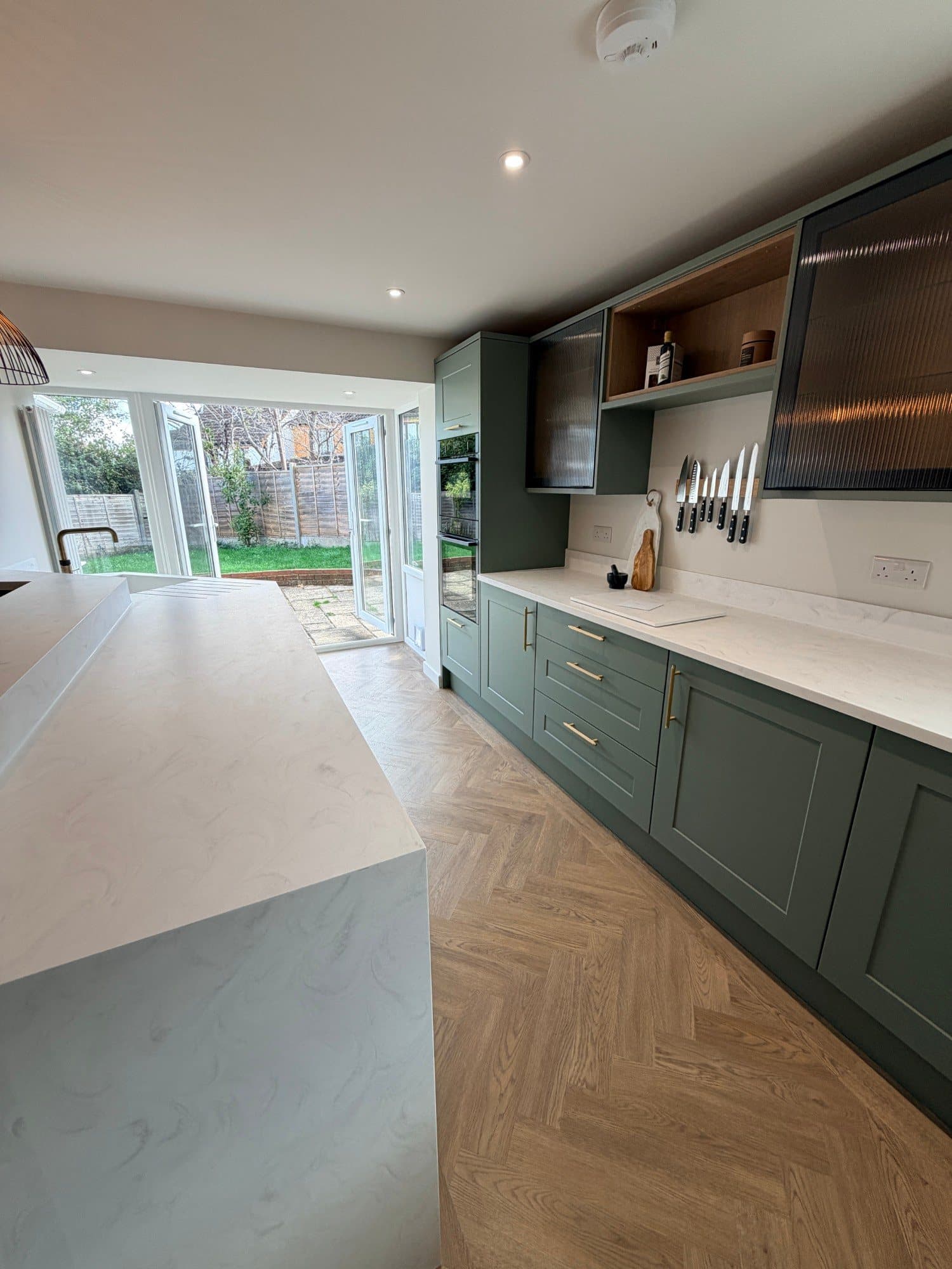 Open-plan sage green kitchen with herringbone flooring and bifold doors onto the garden, Peckover Court, Milton Keynes