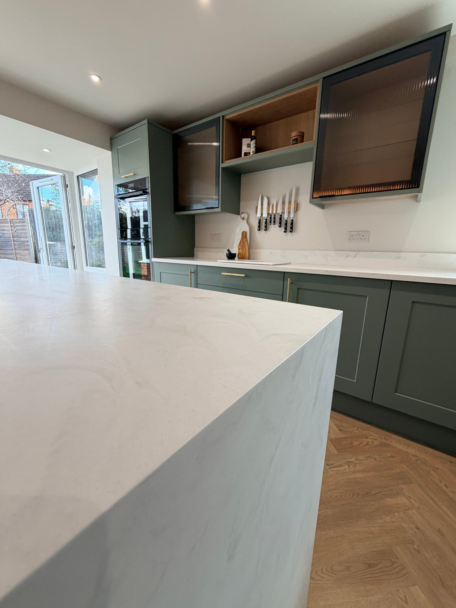 Kitchen island with marble-effect worktop and fitted wall ovens in a sage green kitchen