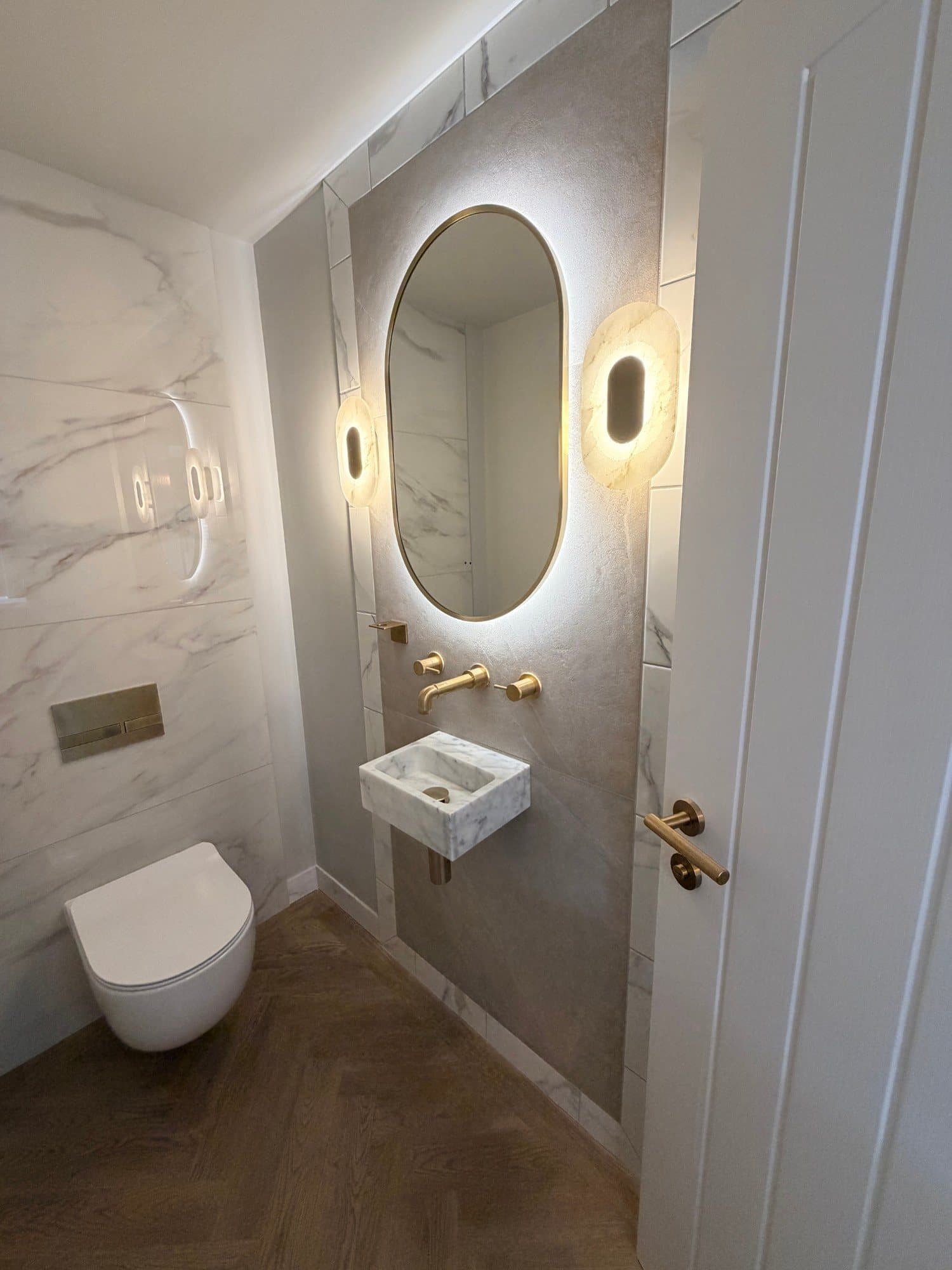Downstairs WC with marble-effect tiling, brass fittings and a backlit oval mirror