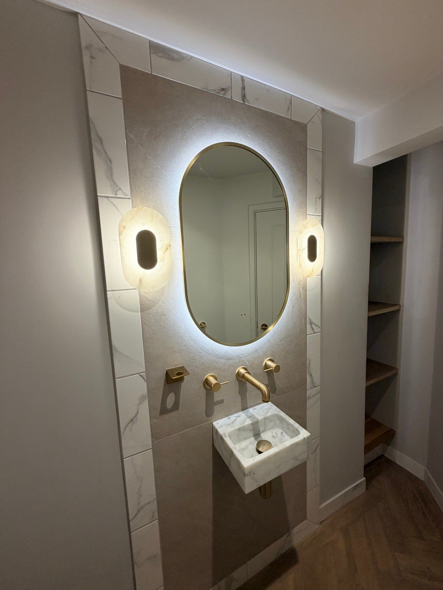 Brass wall-mounted taps and marble basin beneath a backlit mirror