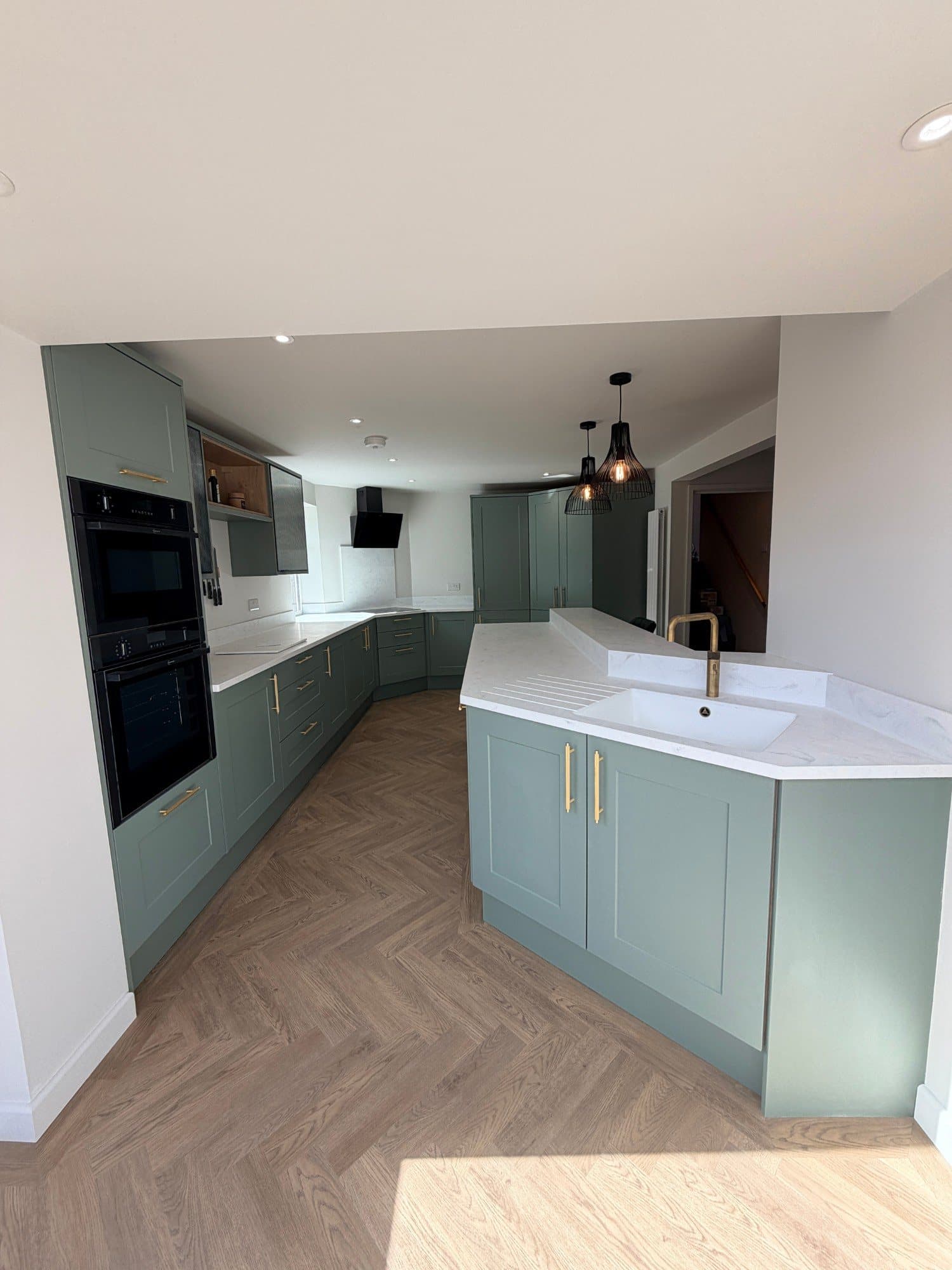 Open-plan sage green kitchen viewed from the doorway, with an island, pendant lighting and herringbone flooring, Peckover Court, Milton Keynes