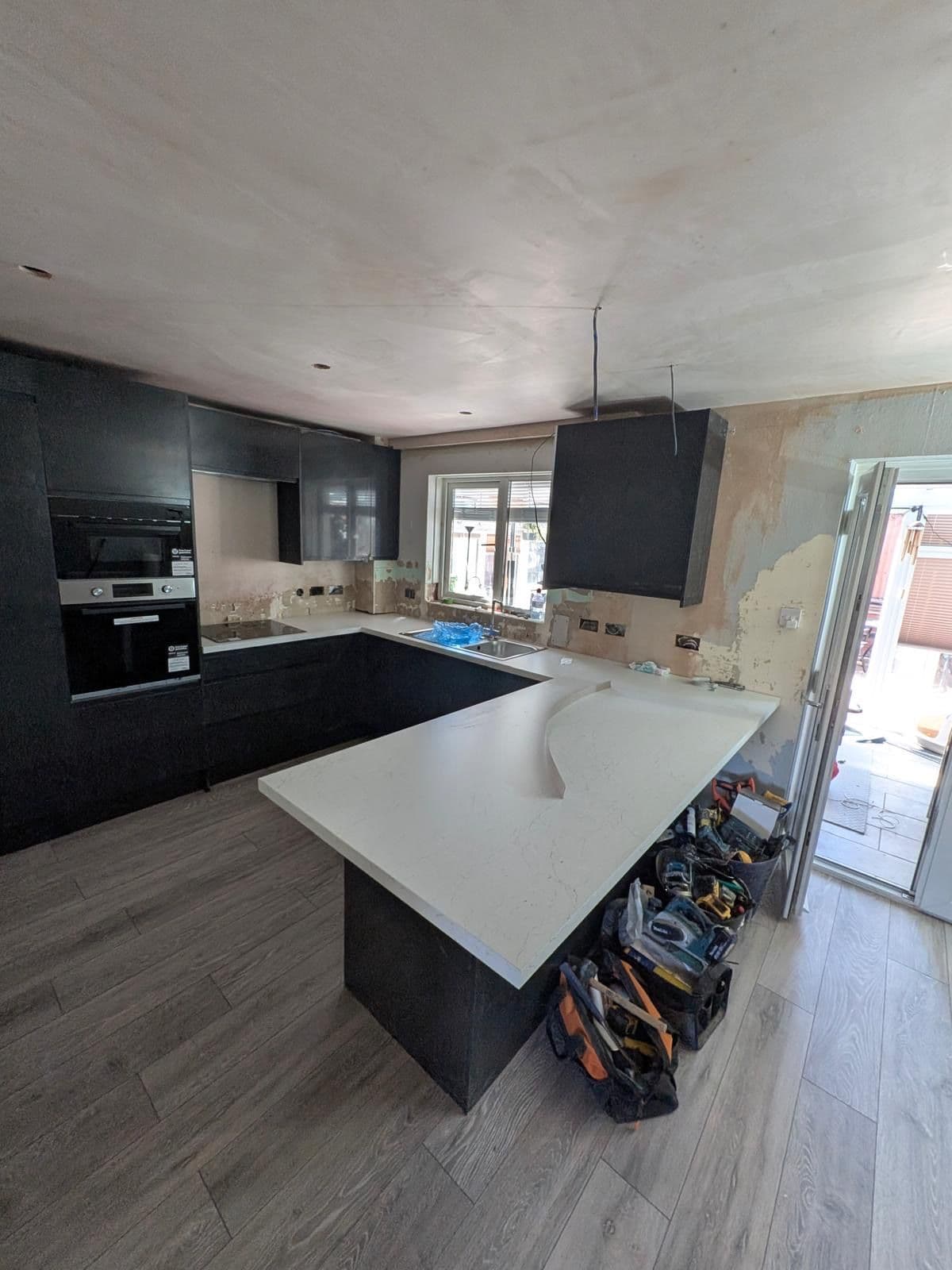 Kitchen under renovation with black cabinetry fitted, white worktop and peninsula, bare plastered walls and exposed wiring