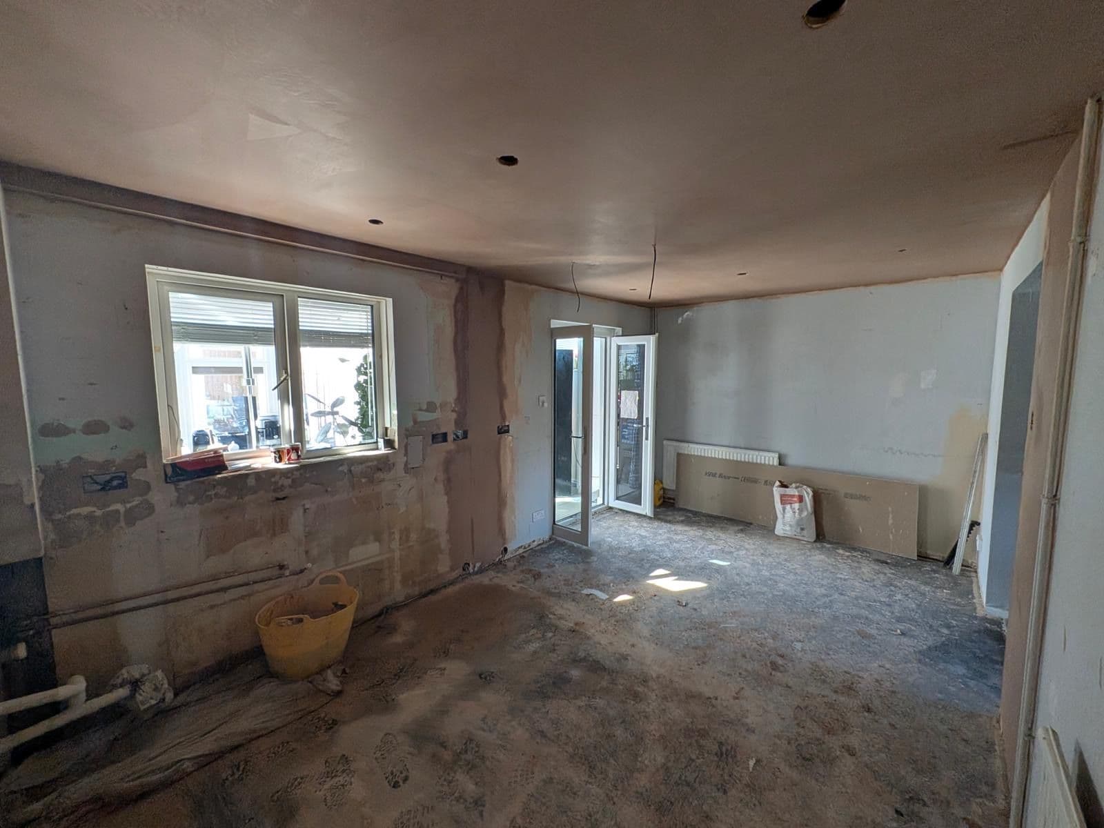 Room stripped back to bare plaster and a concrete floor before renovation, with a window and part glazed door onto the garden