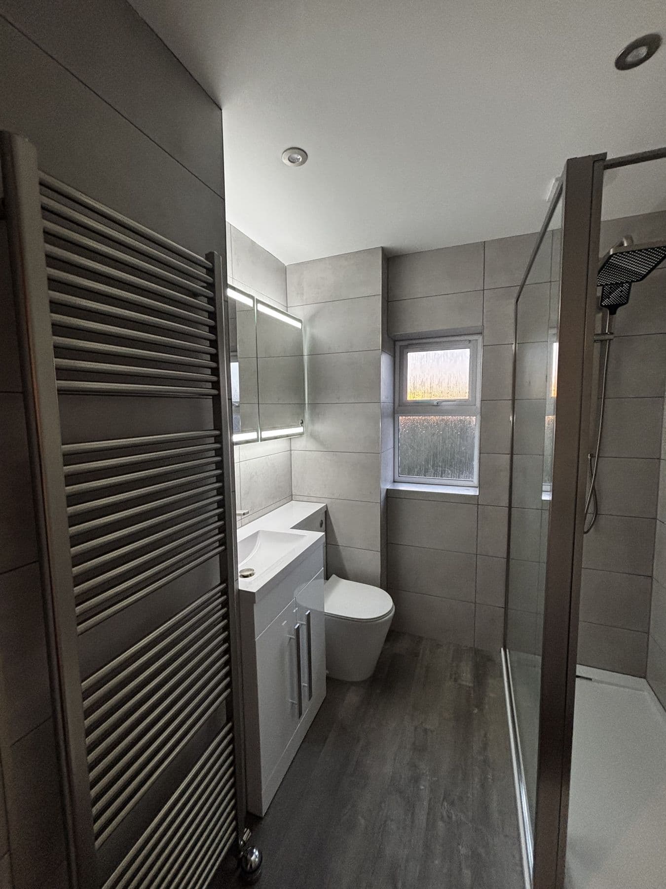 Small bathroom with a chrome ladder towel radiator, mirrored cabinet, white vanity sink and wall hung toilet, and a walk in shower with a glass screen, Playfield Close, Biggleswade