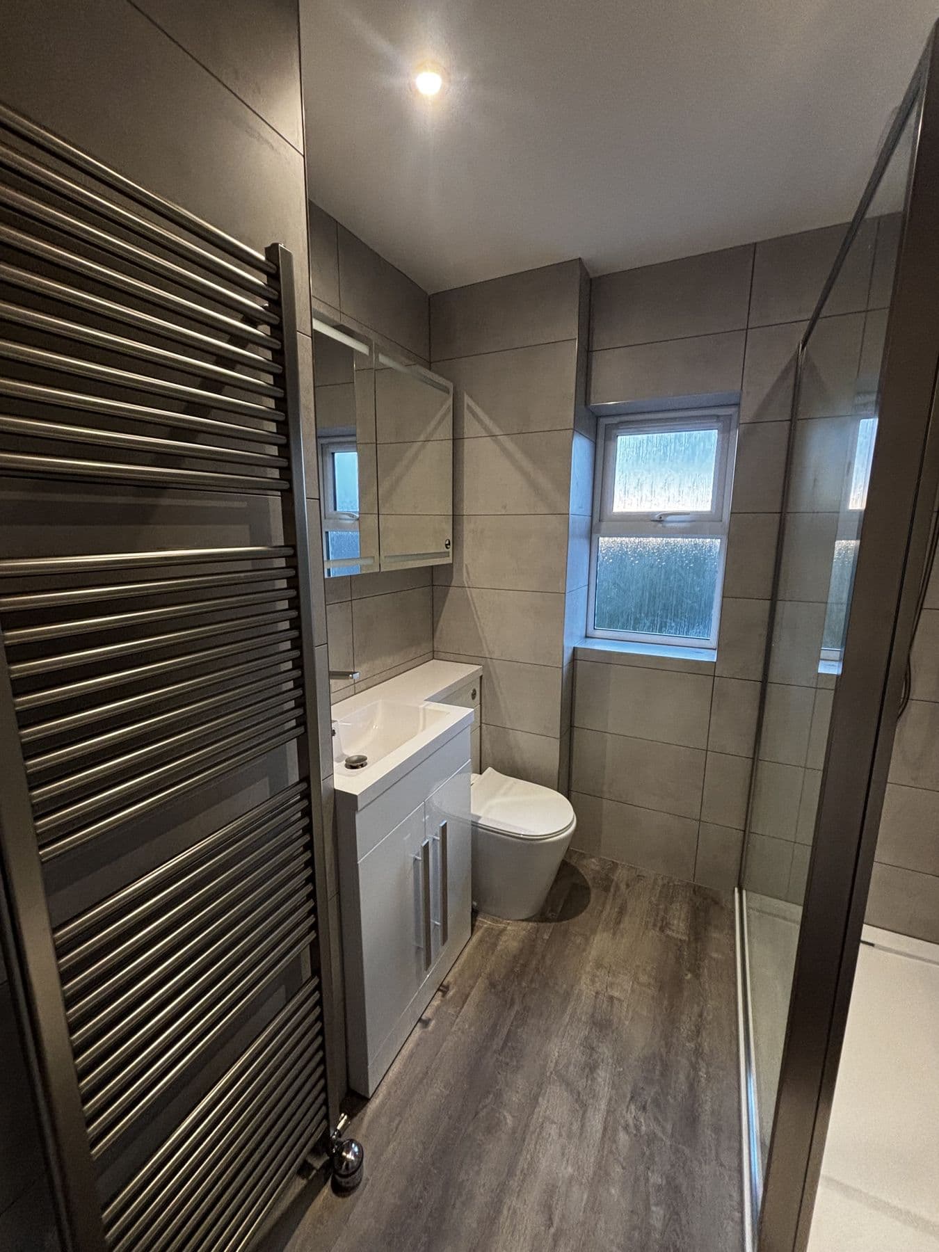 Bathroom with grey tiled walls, a white vanity sink and toilet, a frosted window, and a glass shower screen