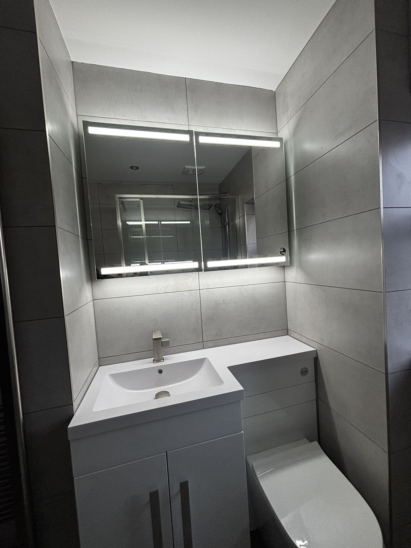 Close up of a white vanity sink under a mirrored cabinet with LED strip lighting, against grey tiled walls
