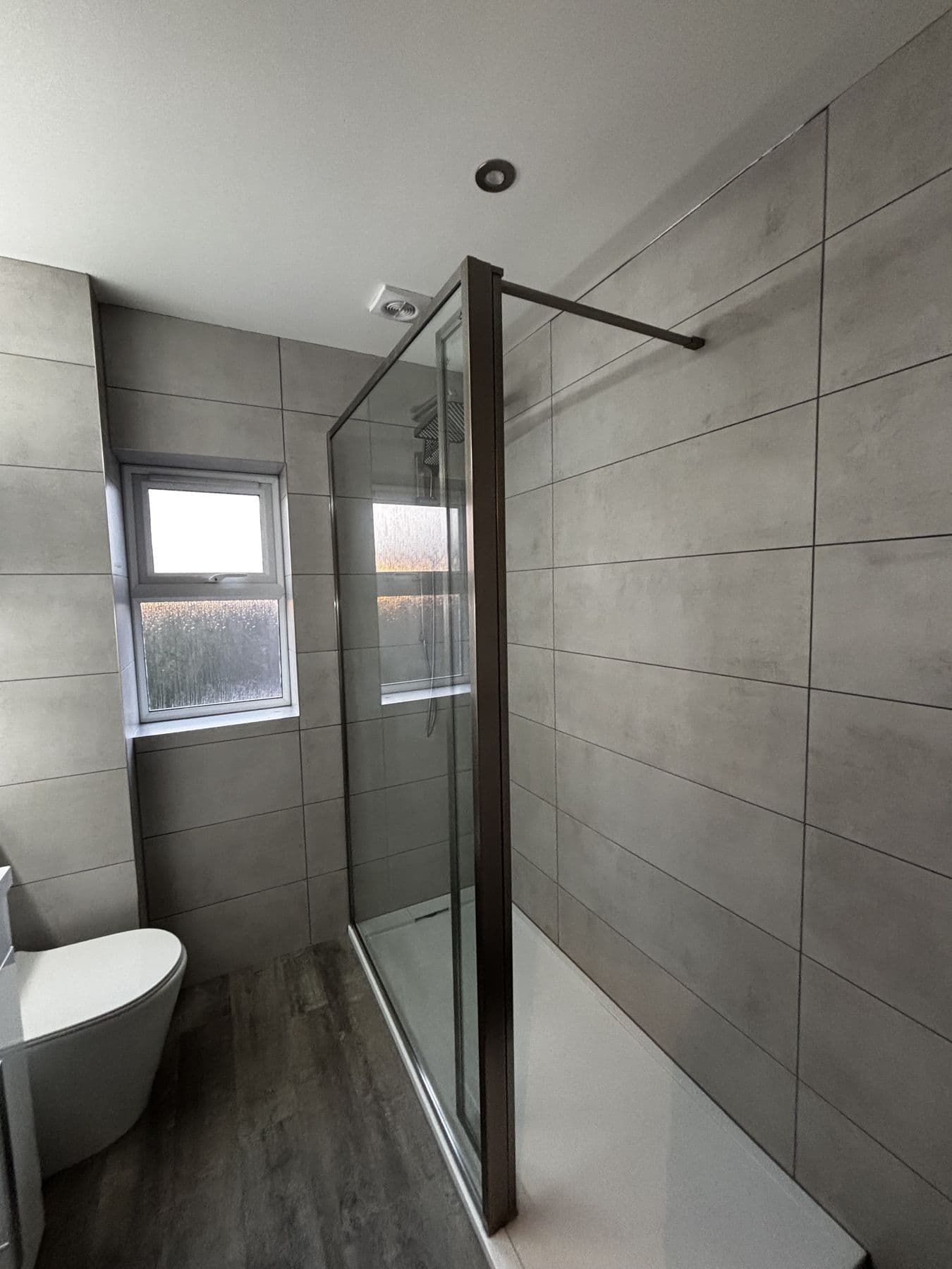 Walk in shower with a frameless glass screen next to a frosted window, grey tiles and dark wood effect flooring