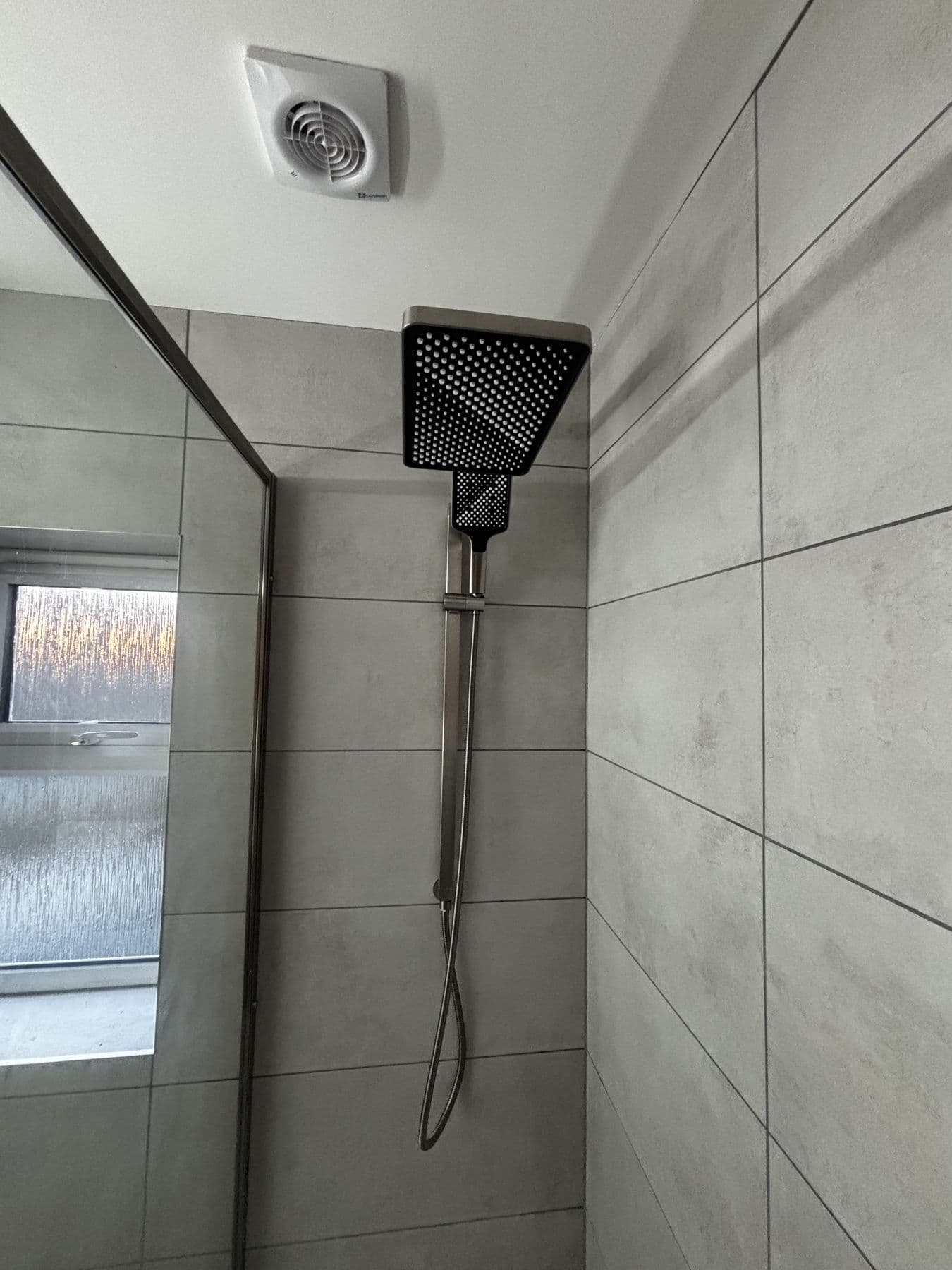 Close up of a rainfall shower head and handheld shower on a riser rail against grey tiles