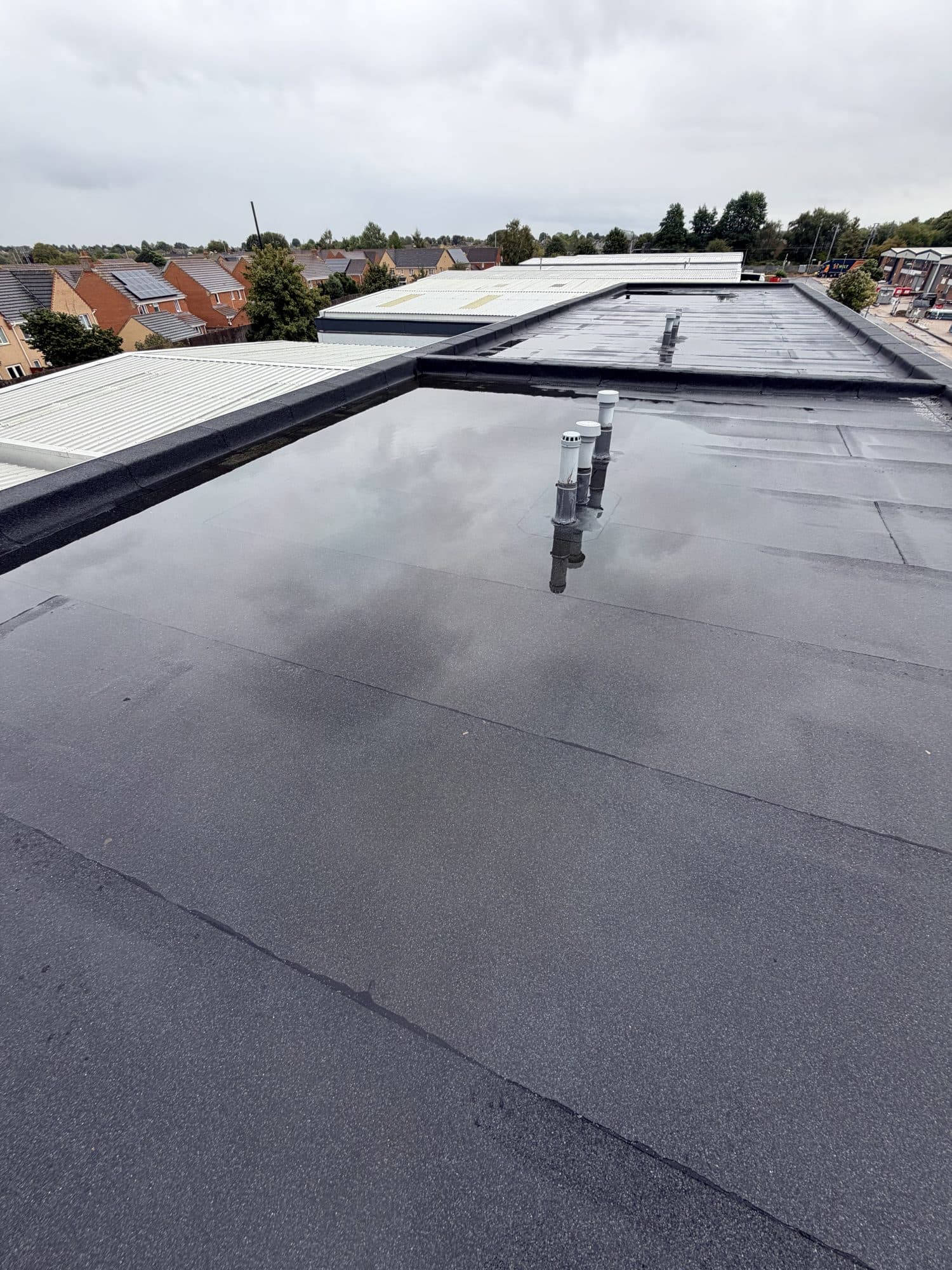Wet flat roof of a commercial unit with three vent pipes, overlooking neighbouring warehouse roofs and housing
