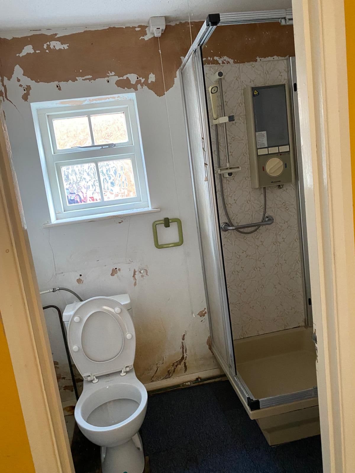 Original bathroom with electric shower unit, peeling walls and stained plaster before renovation